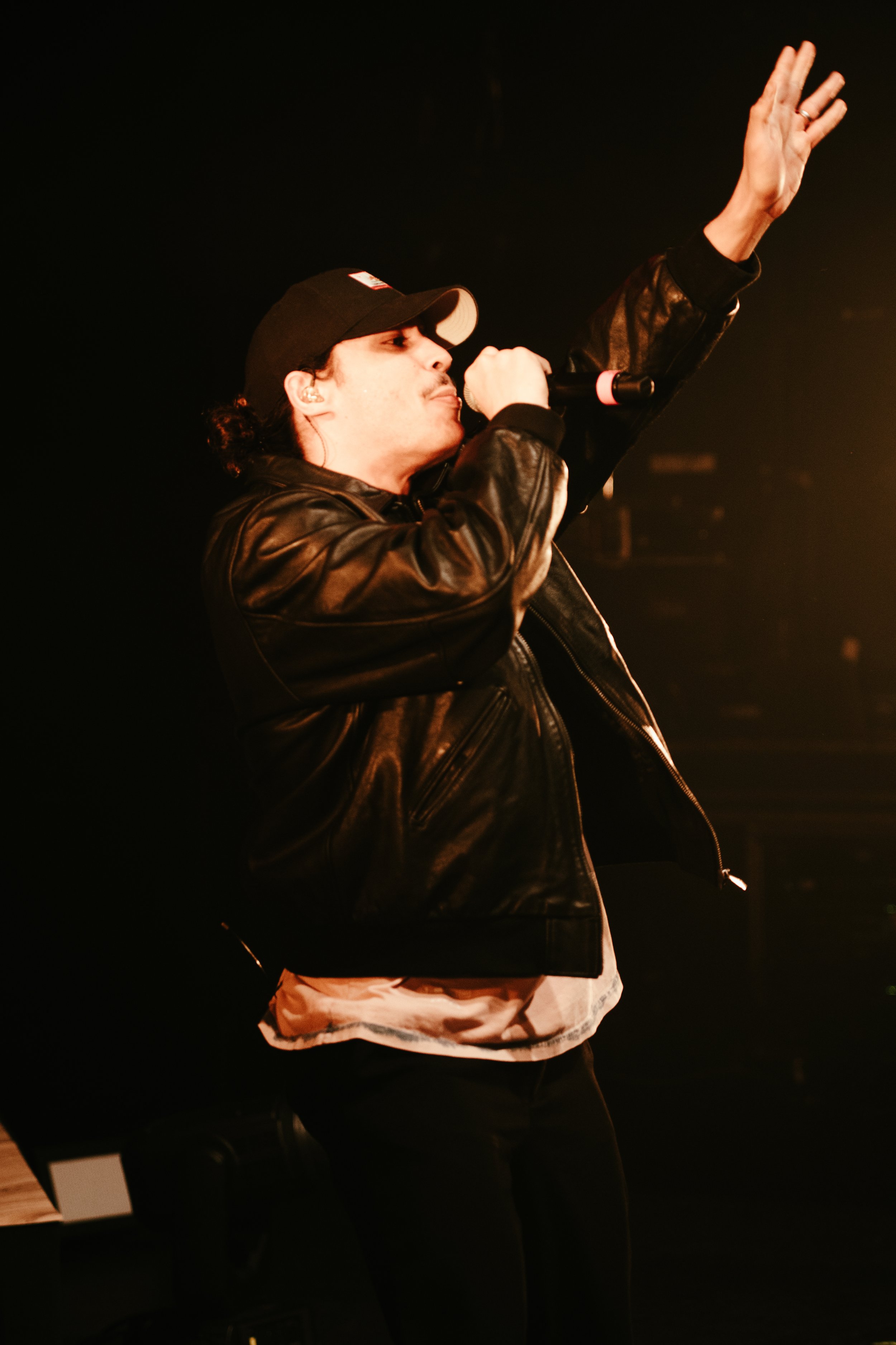 Un chanteur en tenue décontractée, portant une casquette et une veste en cuir, chante dans un micro sur scène, avec un geste expressive de la main.