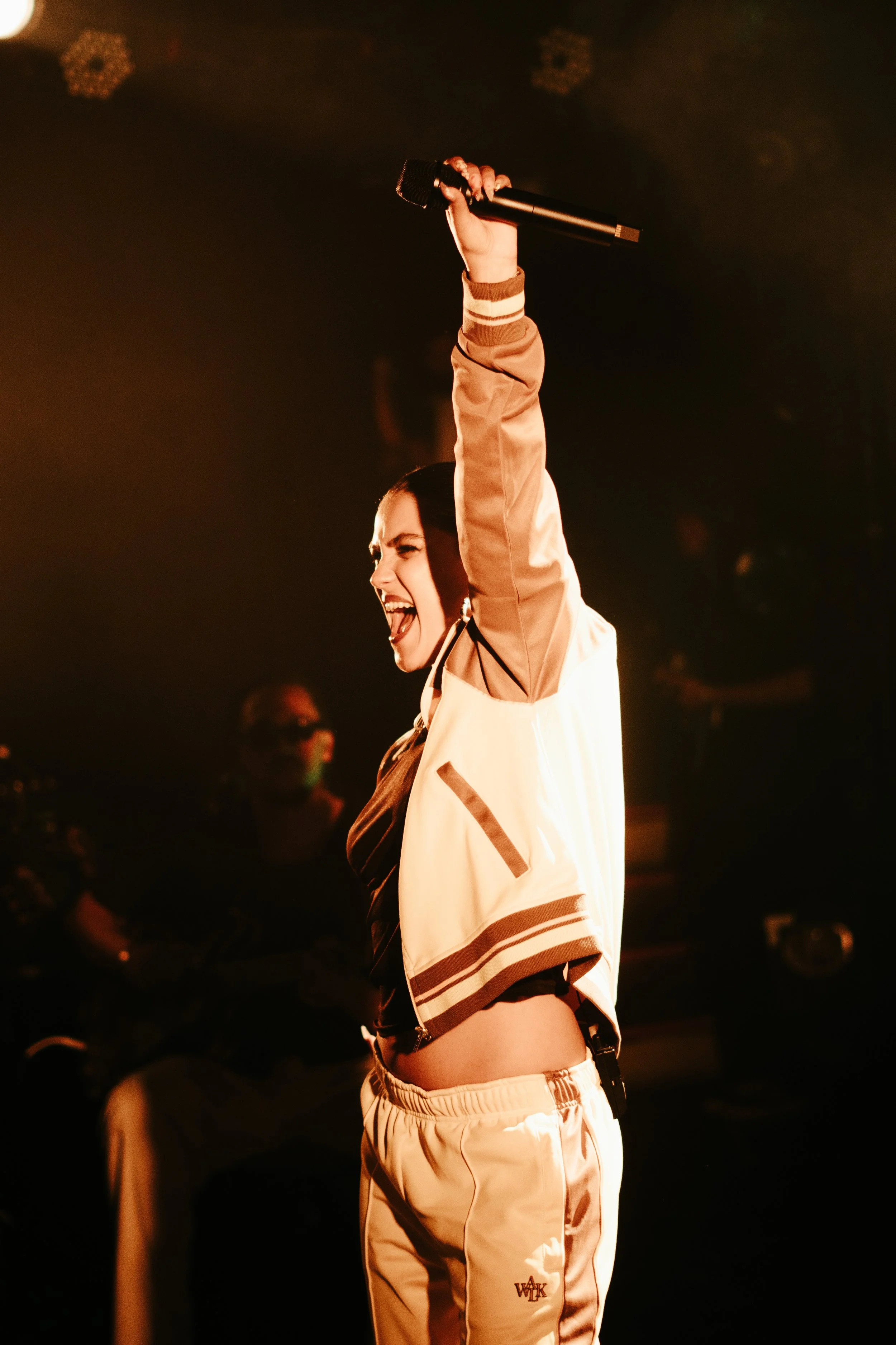 Une femme avec un micro dans la main, criant de joie sur scène, portant un survêtement beige et blanc. Ombres et lumière chaude créent une ambiance énergique.
