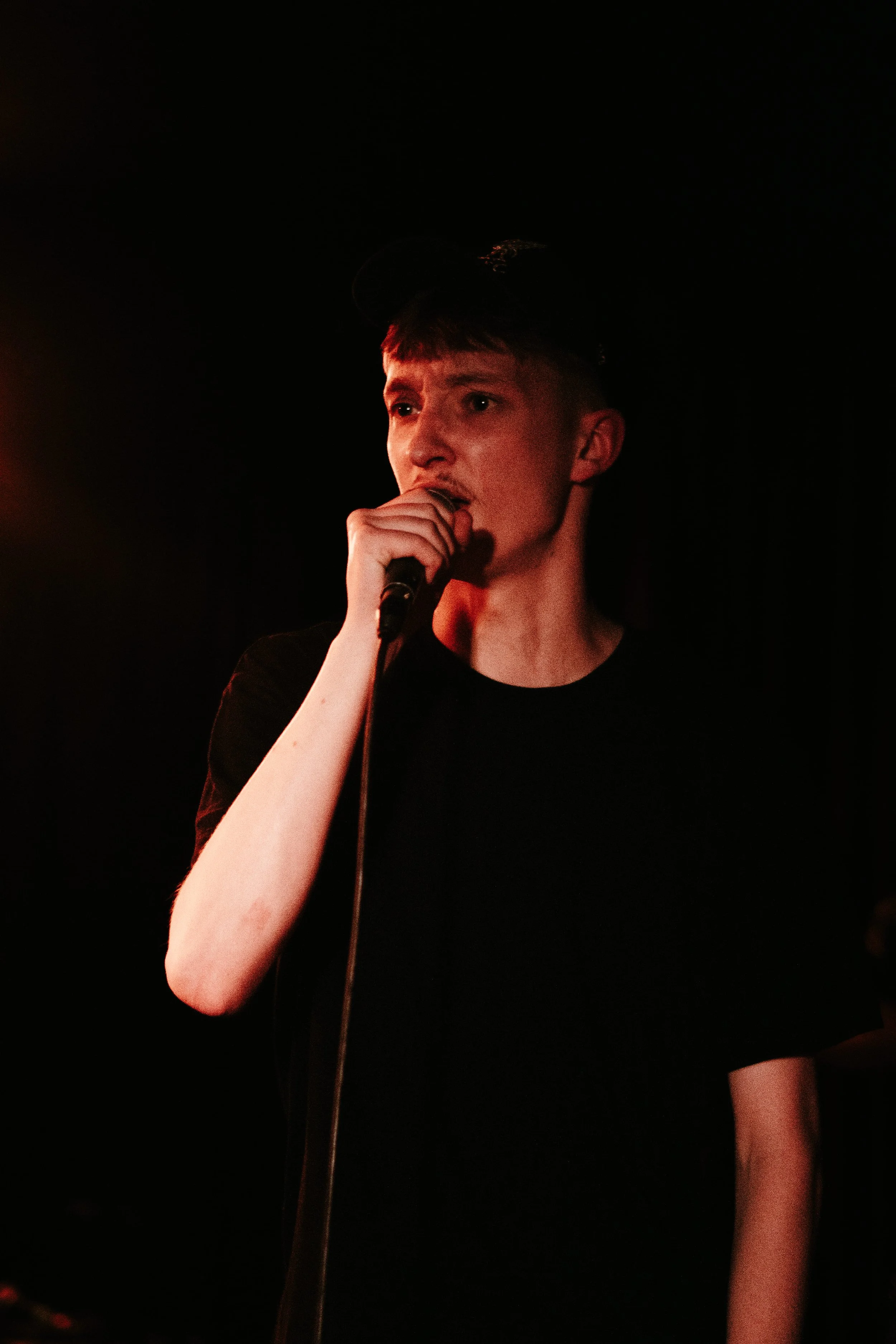 Jeune homme chantant dans une scène sombre, tenant un microphone près de la bouche, portant un t-shirt noir et un bonnet.