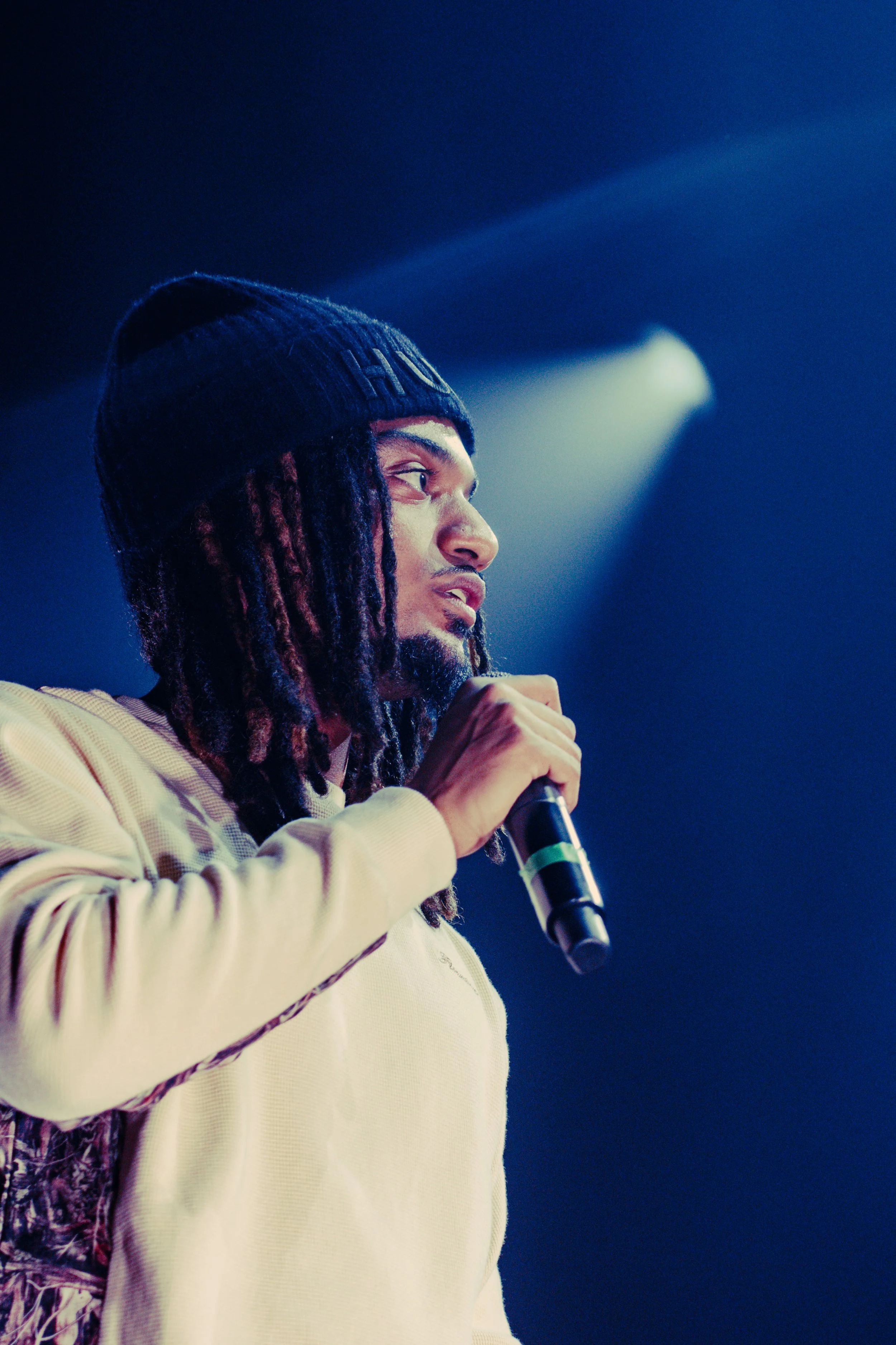 Un homme avec des dreadlocks portant un bonnet noir, tenant un micro lors d'une performance sur scène, éclairé par des lumières bleues.
