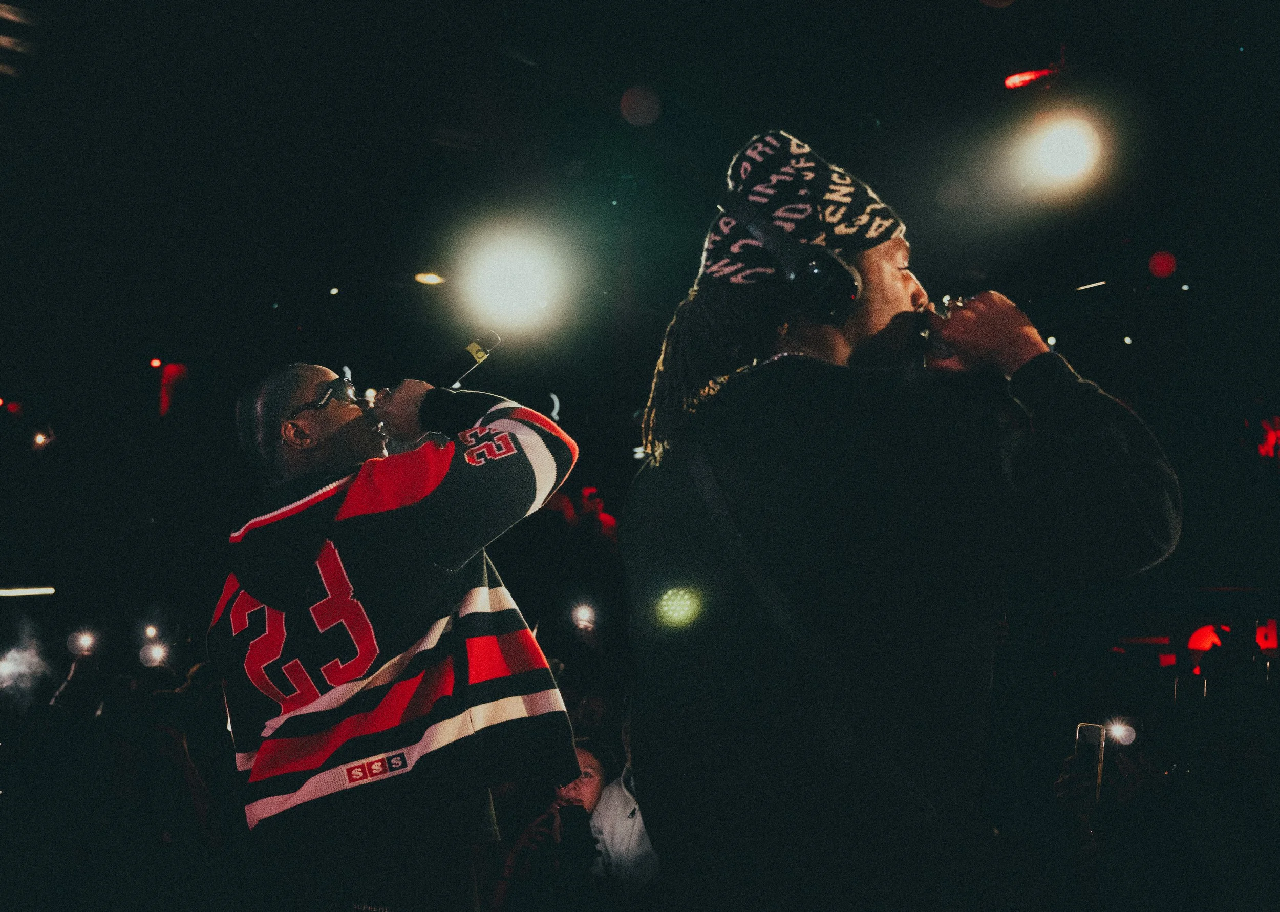 Deux personnes chantant ou parlant dans un micro lors d'une performance ou d'un événement dans une pièce sombre avec un éclairage intense derrière elles.