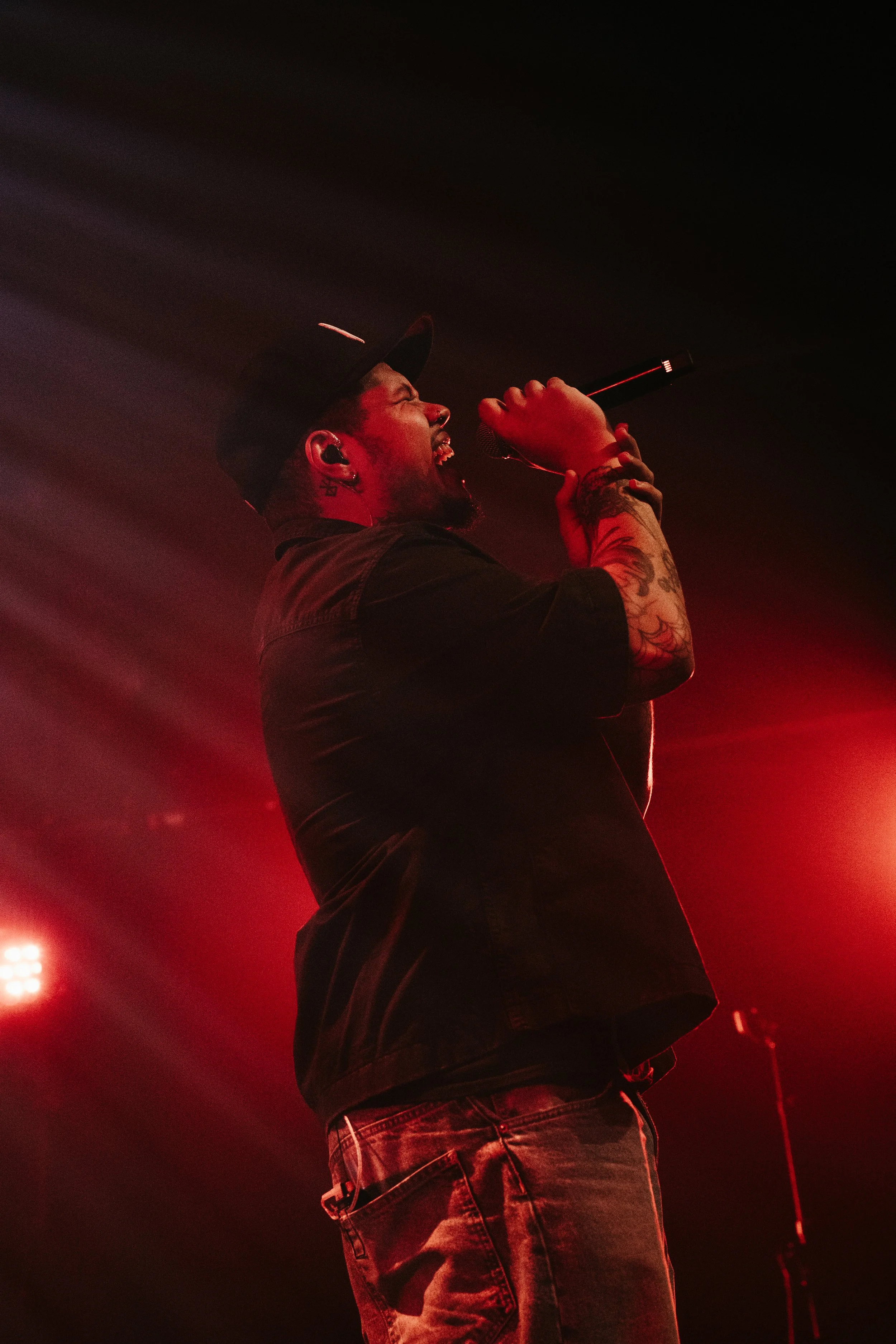 Un homme chantant avec un micro, portant une casquette noire, tatouages visibles sur le bras, dans un environnement sombre éclairé par des lumières rouges.