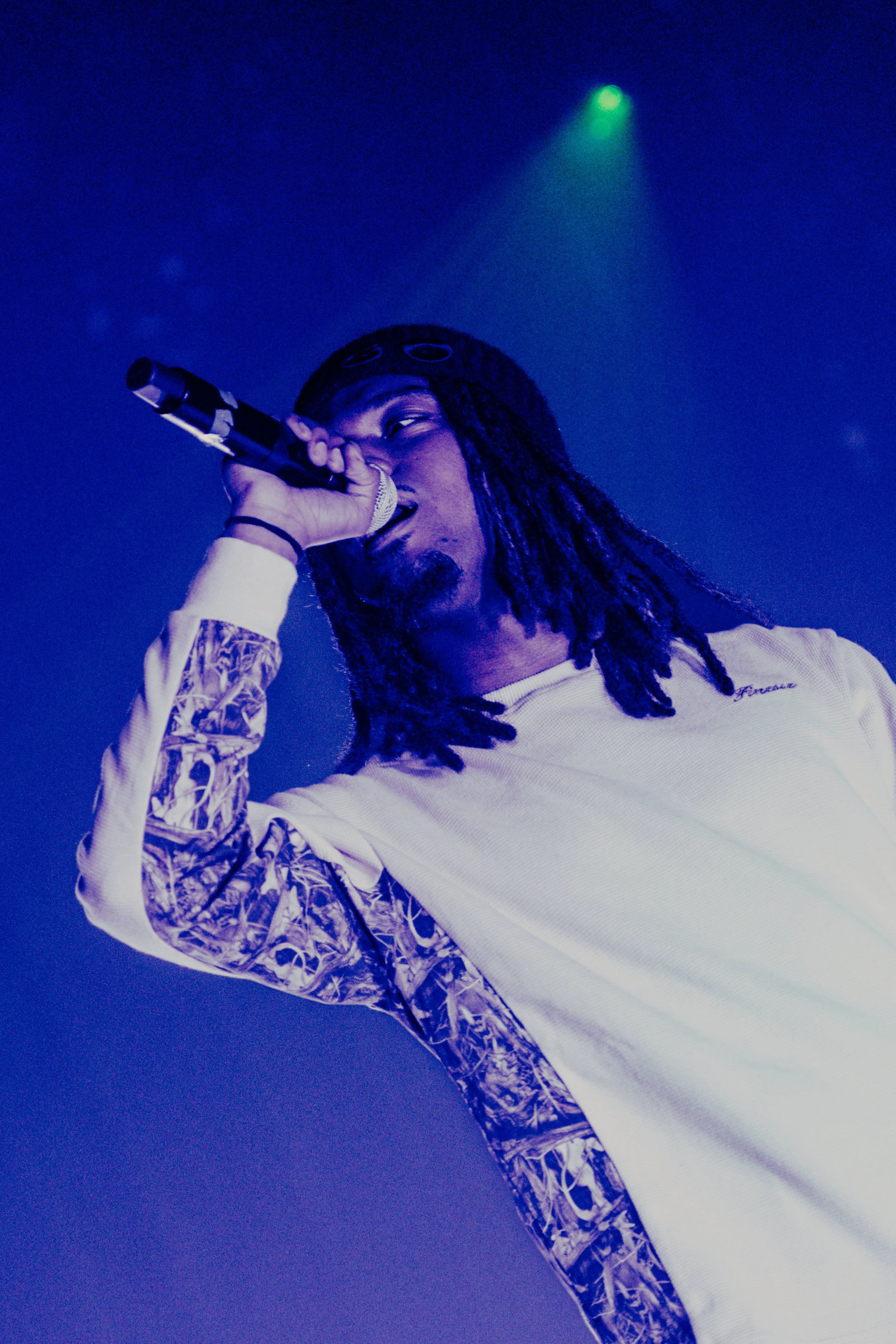 Une personne avec des dreadlocks chante dans un micro, éclairée par des lumières de scène en bleu et vert.