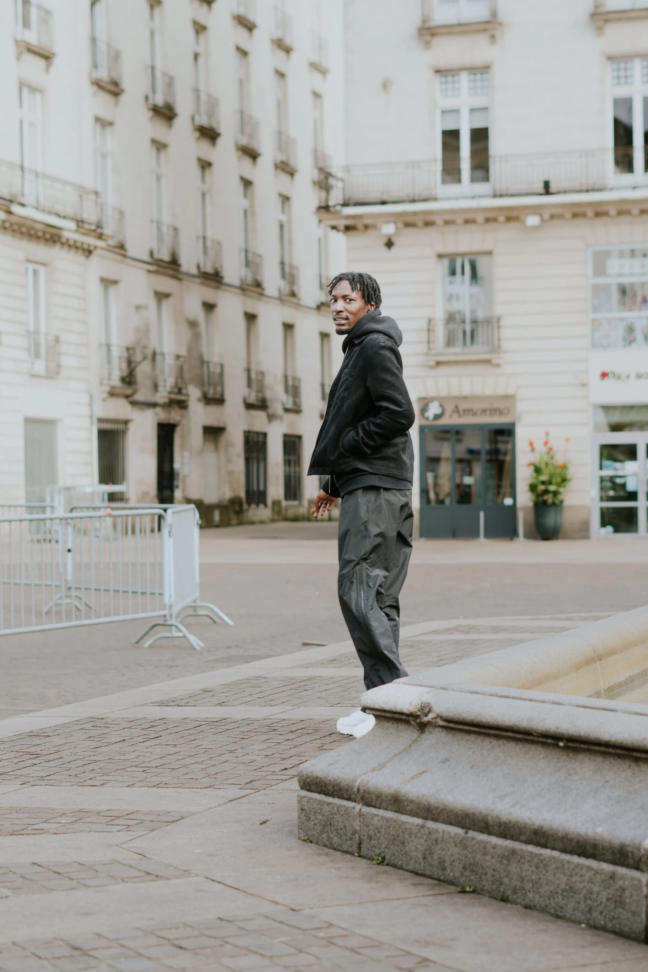 Un homme en vêtement décontracté se tenant sur un trottoir dans une ville avec des bâtiments anciens en arrière-plan.