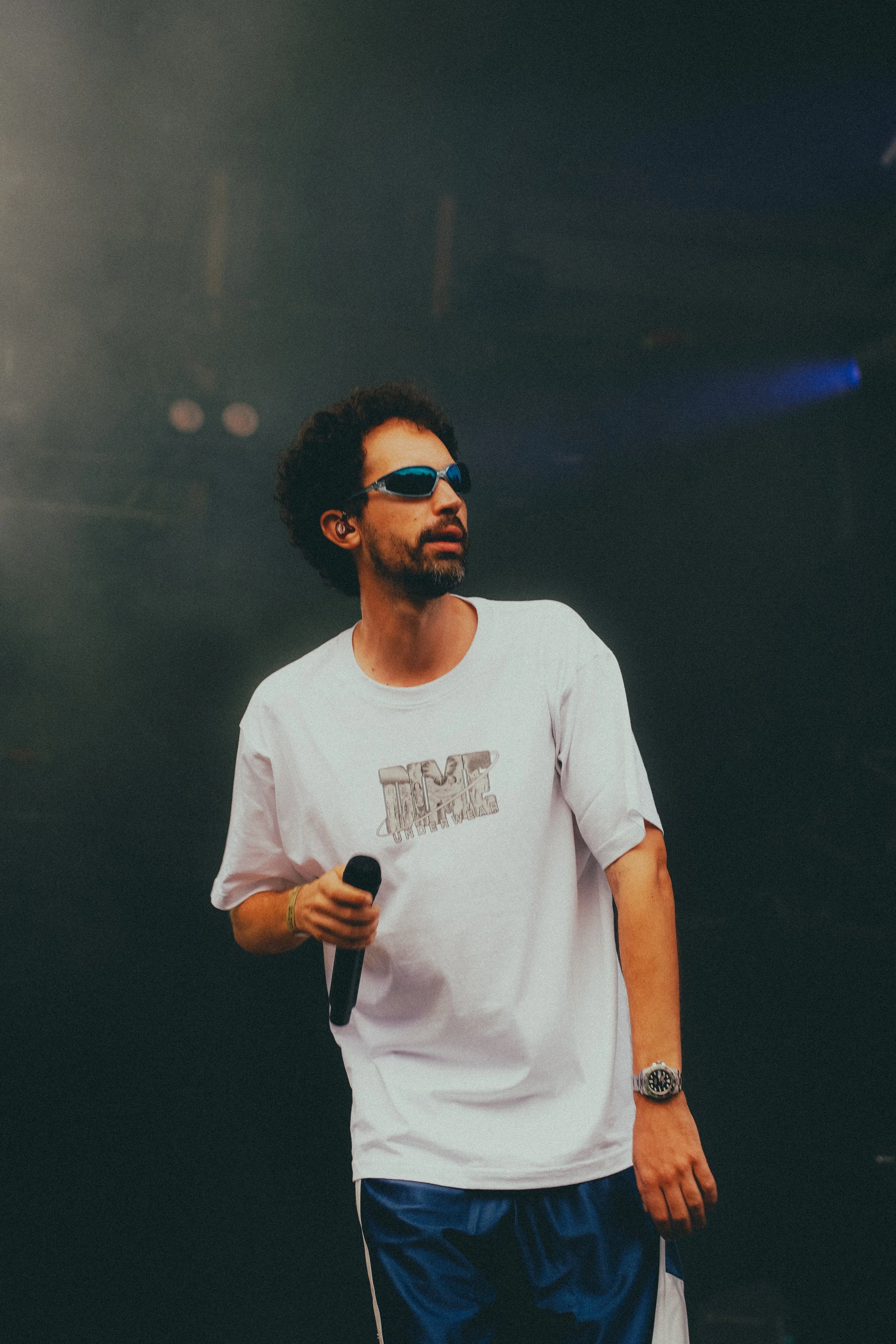 Un homme portant des lunettes de soleil noires, une chemise blanche avec un logo, et un pantalon bleu, tenant un microphone dans la main, semble être en train de se produire sur scène