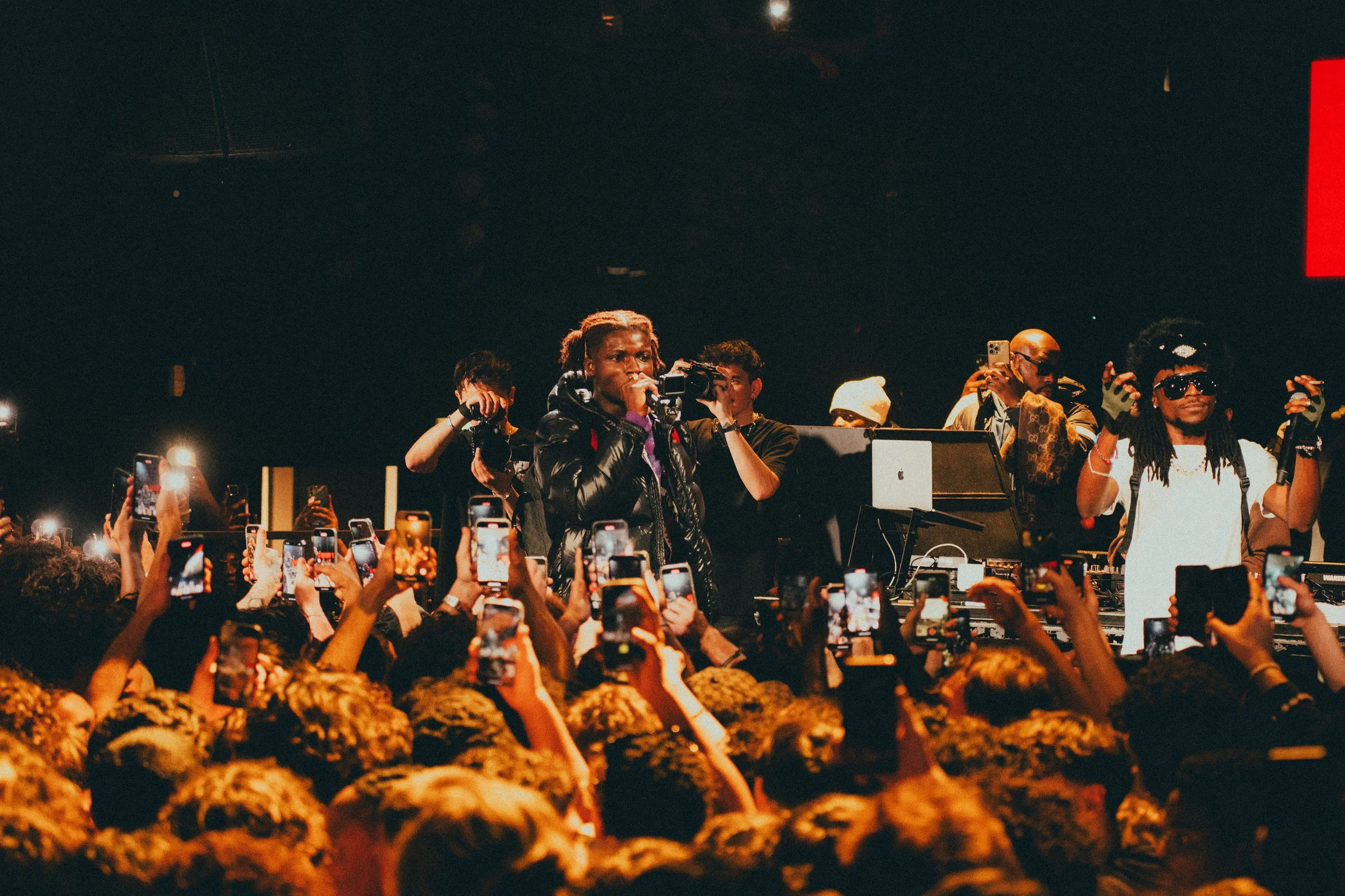 Un concert ou une performance musicale avec une foule qui prend des photos, plusieurs artistes sur scène, dont un homme avec des dreadlocks, un autre avec une selle cap, et un DJ en arrière-plan.