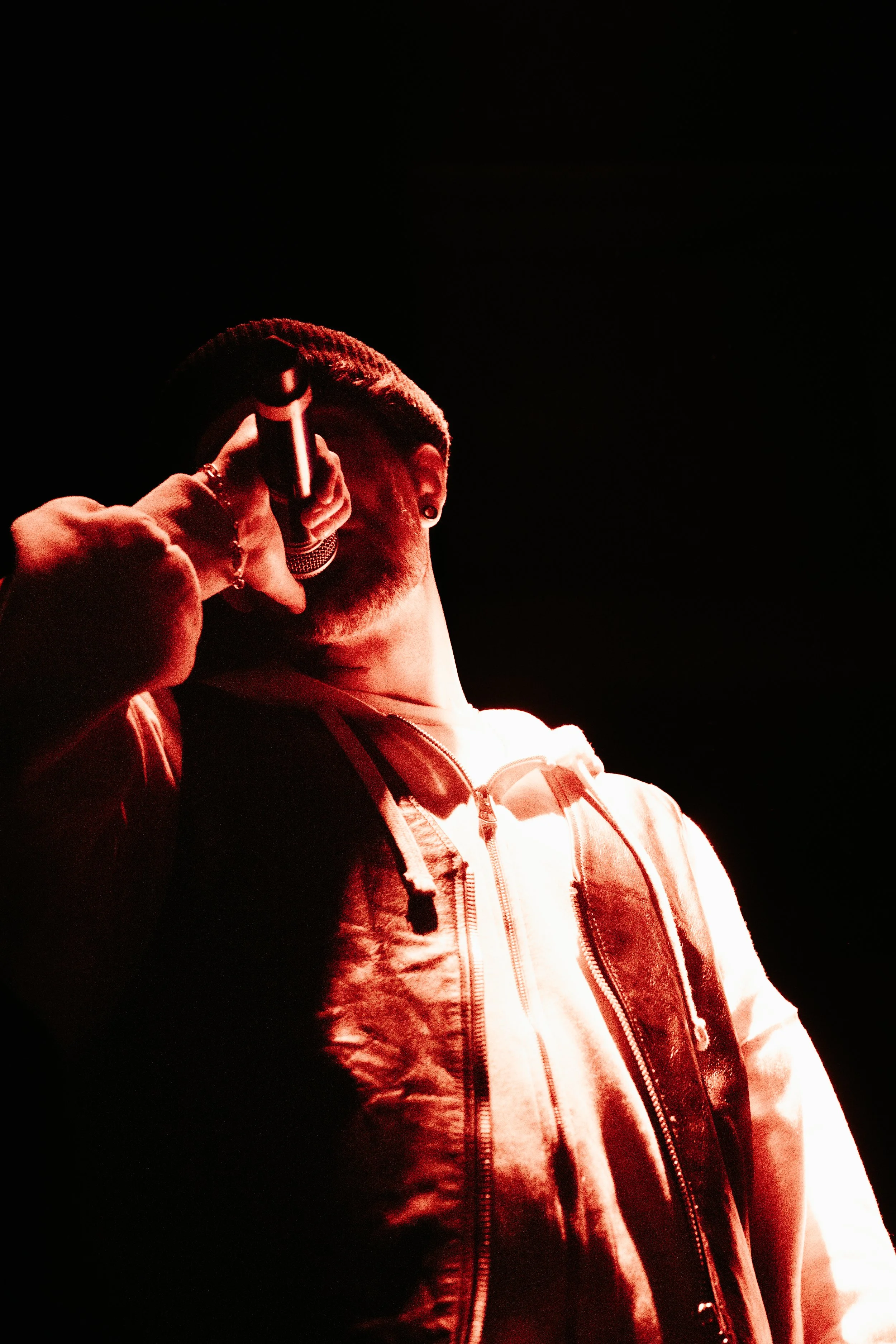 Un jeune homme tenant un microphone à la main dans un éclairage rouge, portant une veste à capuche et un t-shirt blanc, dans un environnement sombre.