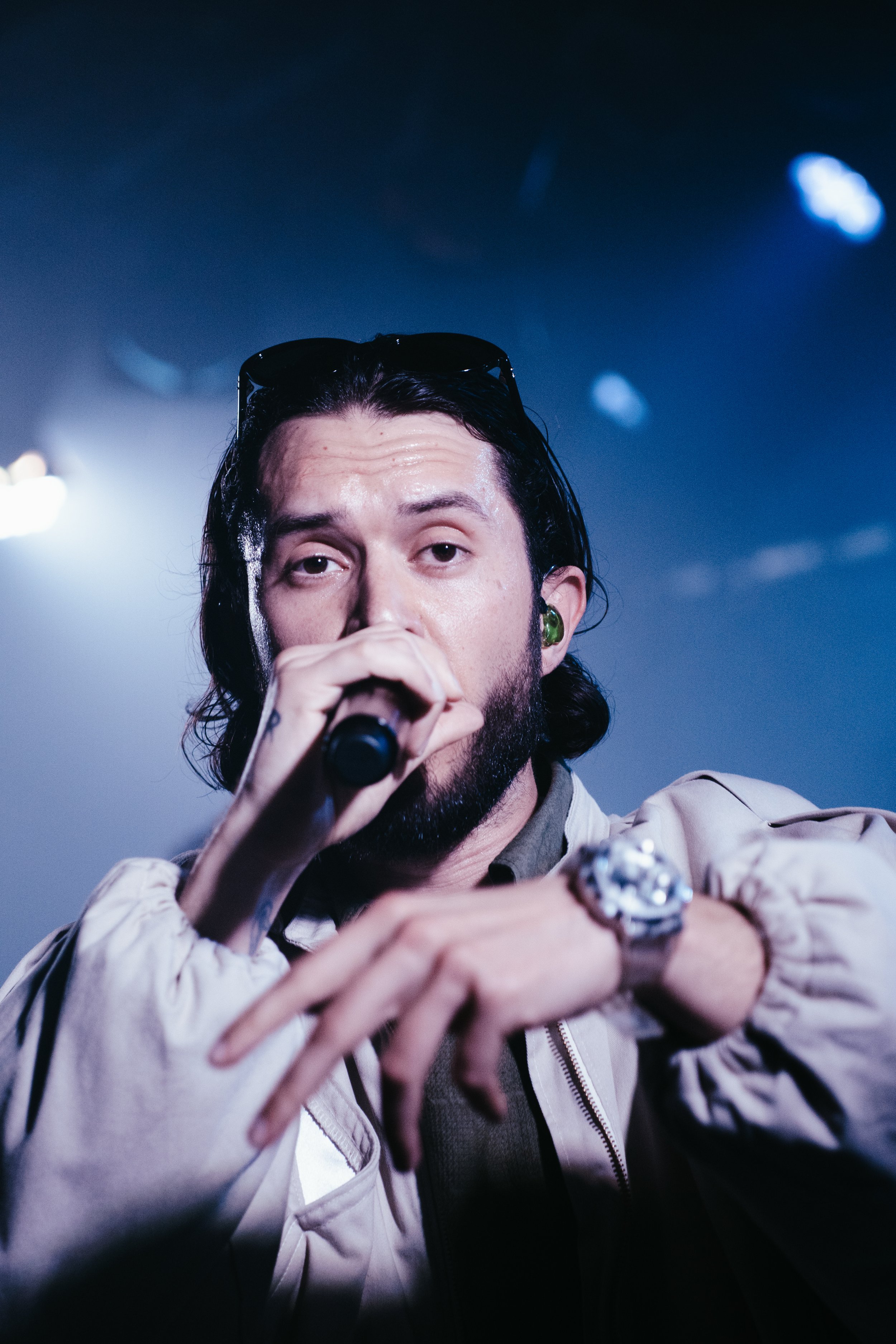 Un homme avec un long cheveux noirs, portant des lunettes noires sur la tête, un peu de maquillage sur le visage, tenant un micro et regardant devant lui, lors d'une performance sur scène avec un fond bleu.