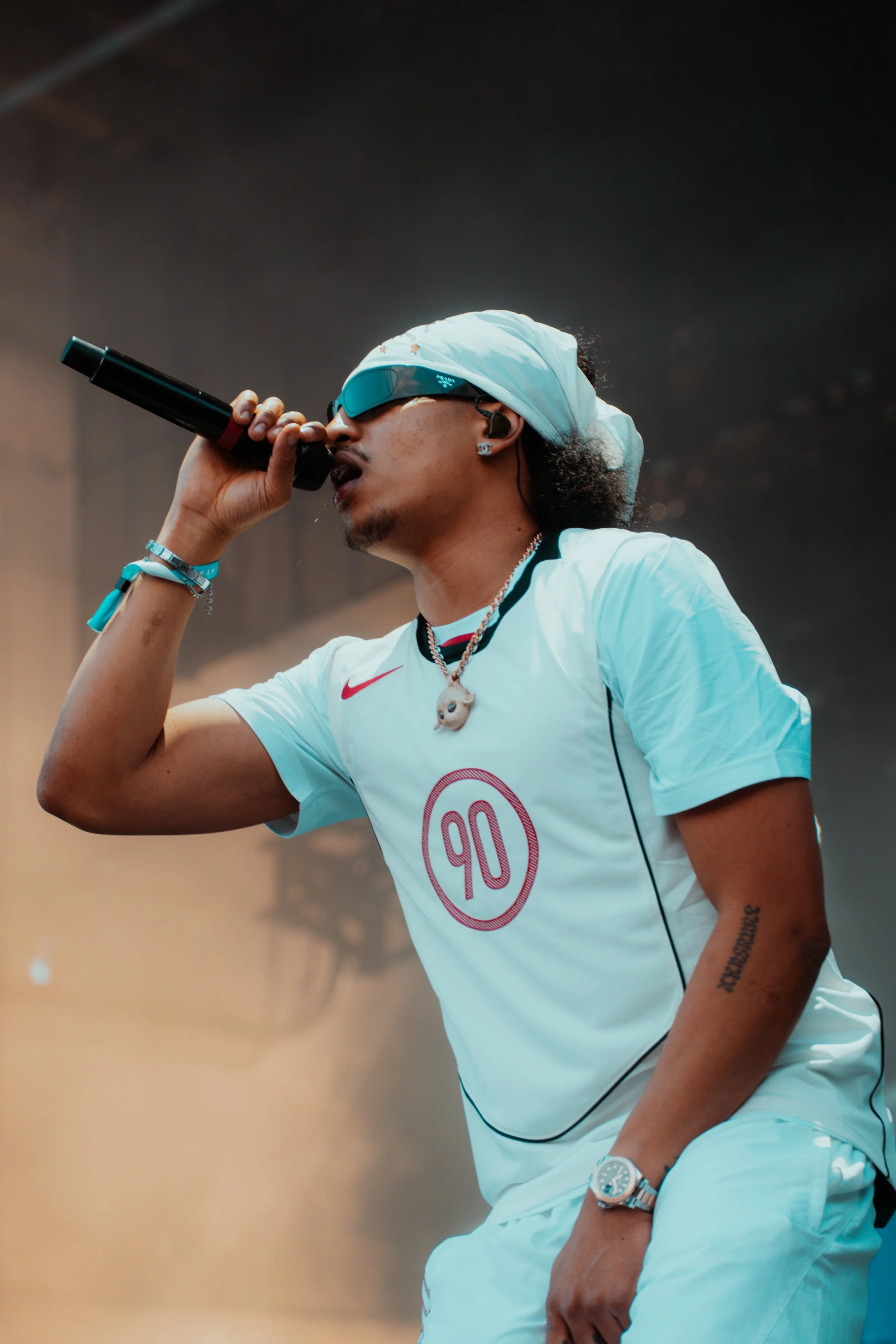 Un homme avec des lunettes de soleil et un bandana blanc chante dans un microphone, portant un maillot de sport blanc avec un logo et des accessoires.