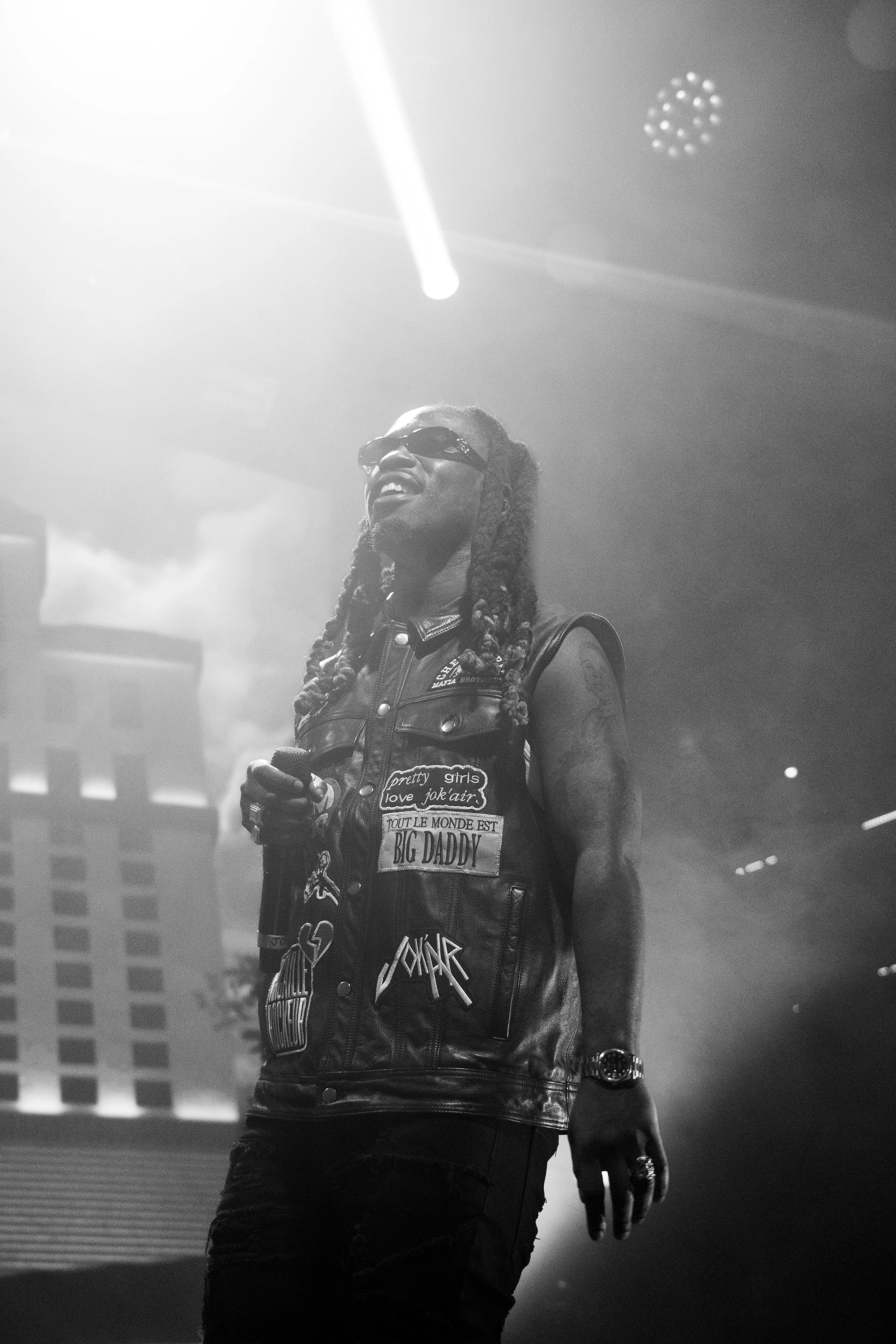 Un homme avec des dreadlocks portant des lunettes de soleil, un gilet en cuir avec des patches, tenant un micro, lors d'une performance en concert.