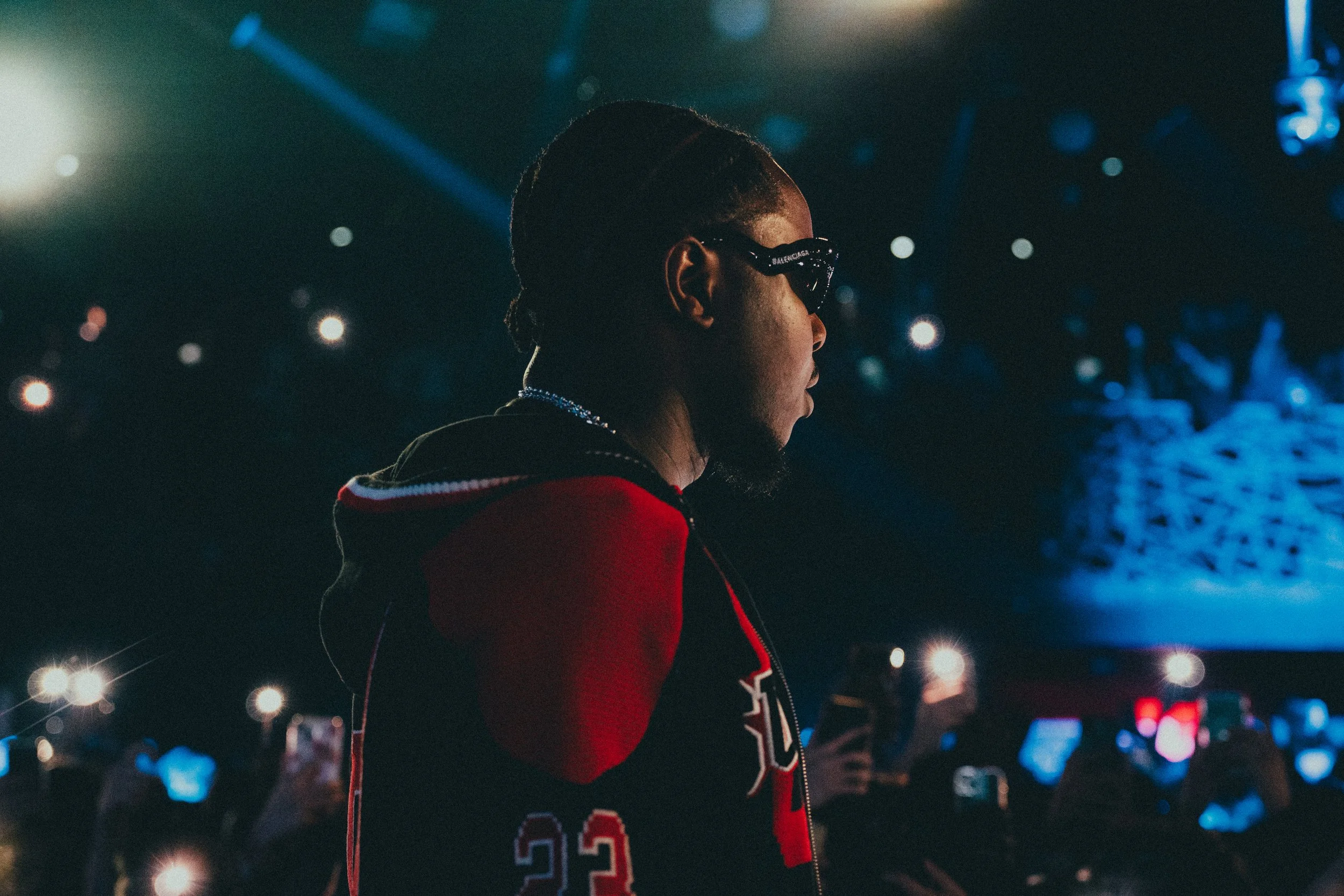Un homme portant des lunettes de soleil, une veste à capuche rouge et noire, dans une ambiance sombre avec des lumières de scène, entouré de personnes utilisant leur téléphone pour prendre des photos.
