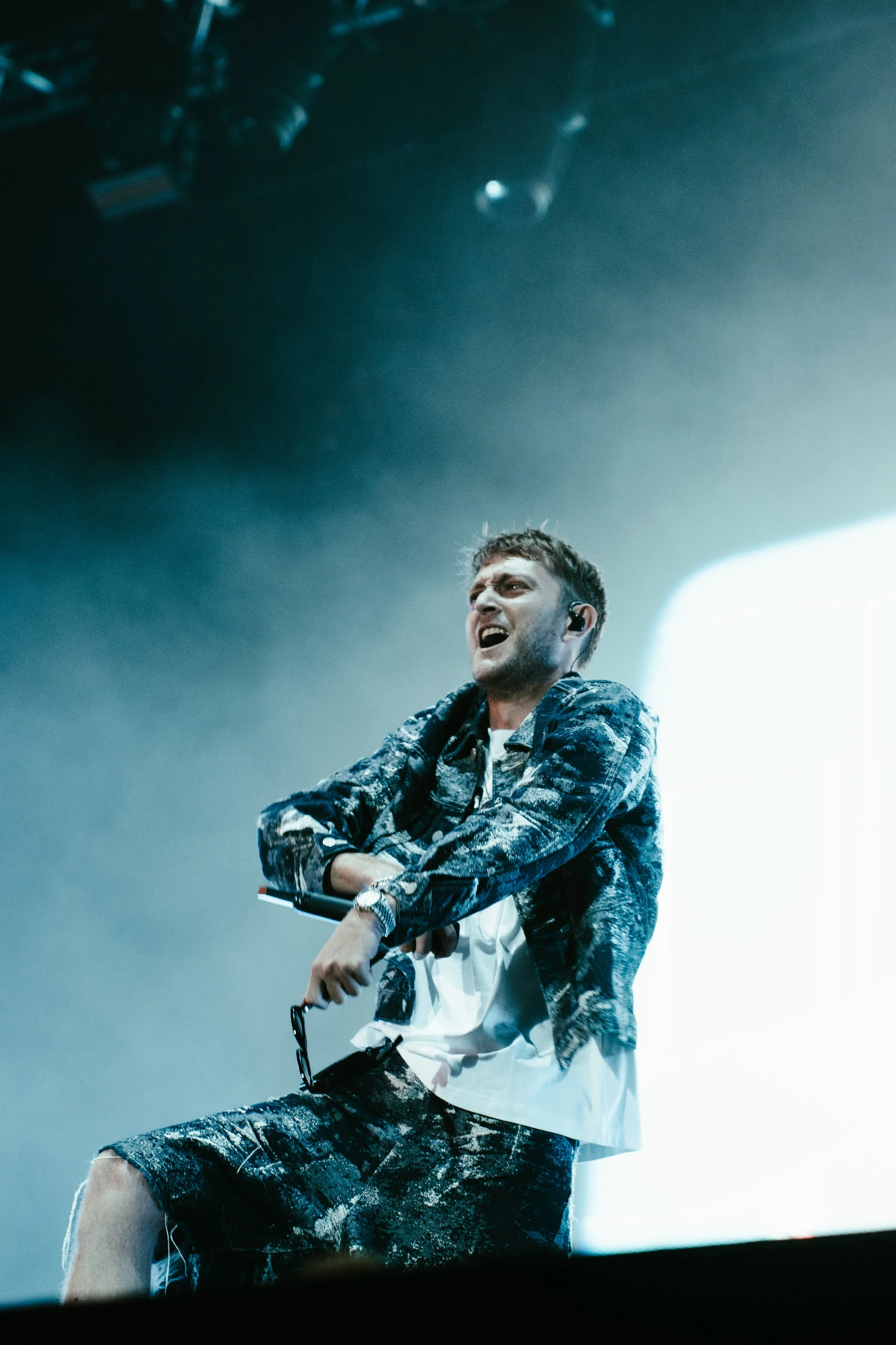 Un homme en tenue décontractée sur scène, visiblement en train de chanter ou de parler, avec un micro à la main, sous un éclairage de scène.