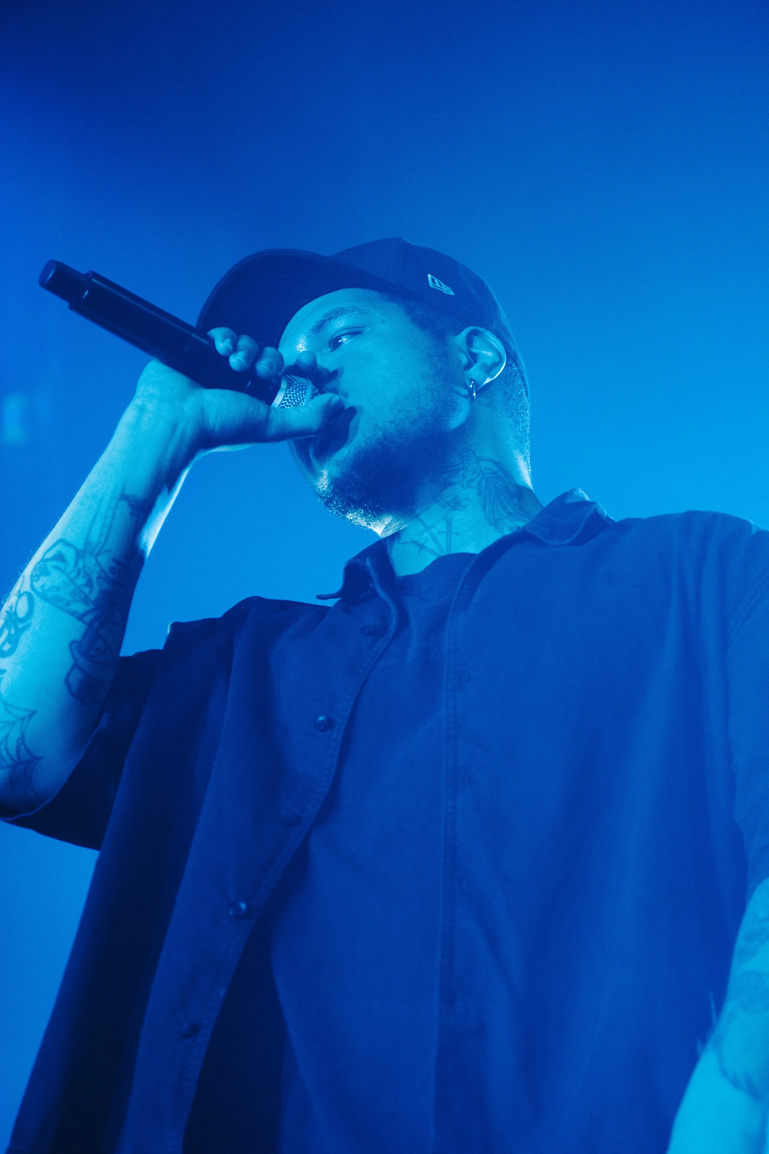 Un homme portant une casquette noire, avec des tatouages visibles sur le bras et le cou, chanter ou parler dans un microphone sous un éclairage bleu.