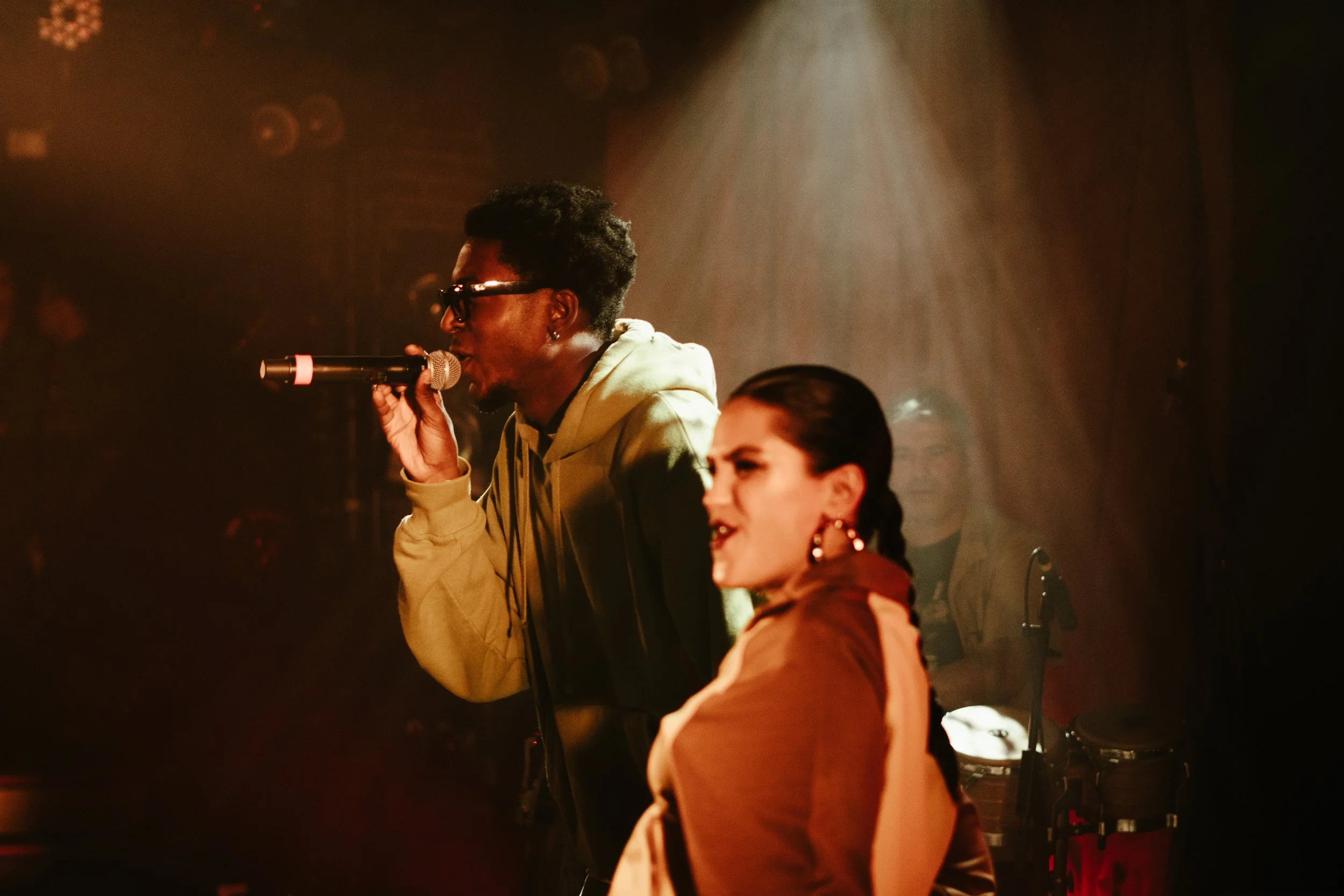 Deux personnes, un homme noir portant des lunettes et un hoodie, chante en utilisant un microphone, et une femme avec des boucles d'oreilles et une veste marron, semble chanter ou parler, dans une scène de performance avec un éclairage chaud et ambia