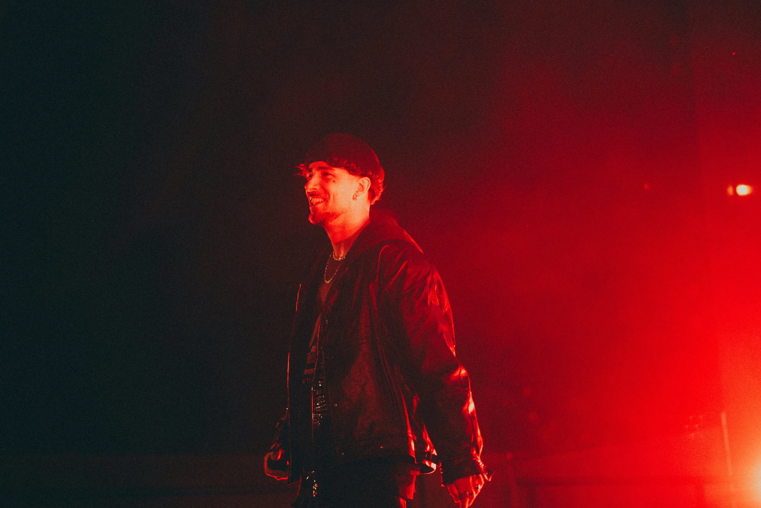 Un homme souriant avec des cheveux noirs courts, portant une veste en cuir noir, sous un éclairage rouge intense.