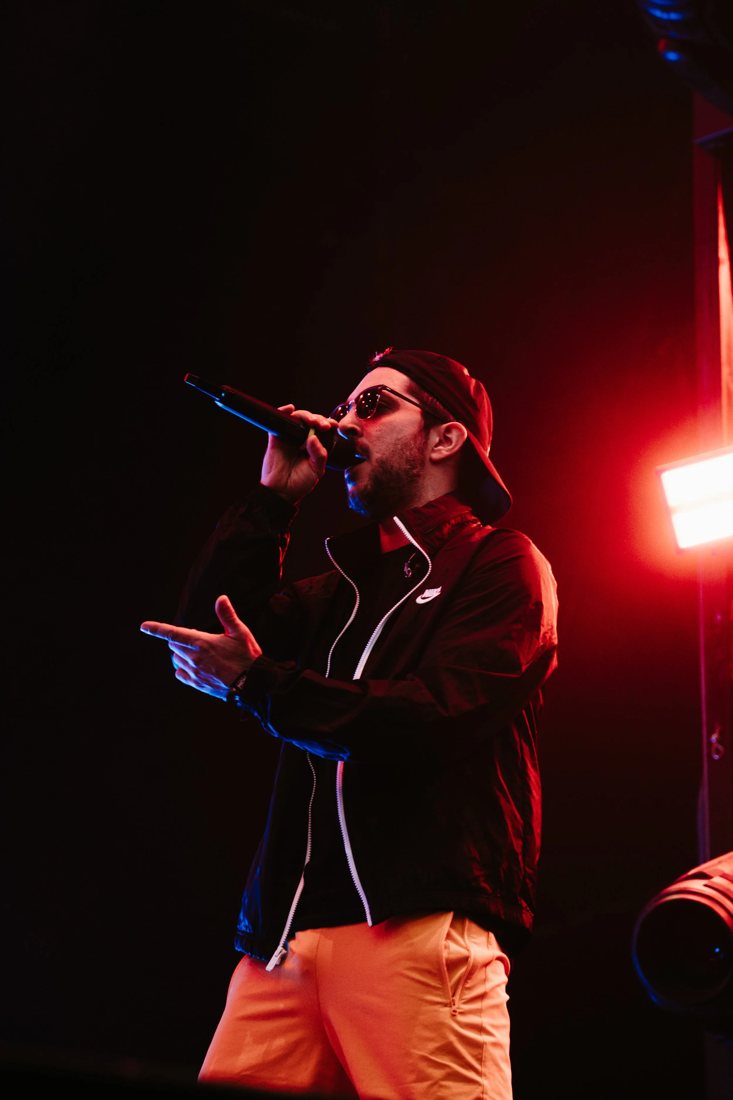 Jeune homme portant lunettes de soleil, chapeau à l'envers, veste Nike, pantalon orange, chantant avec microphone dans un environnement sombre éclairé par des lumières rouges.