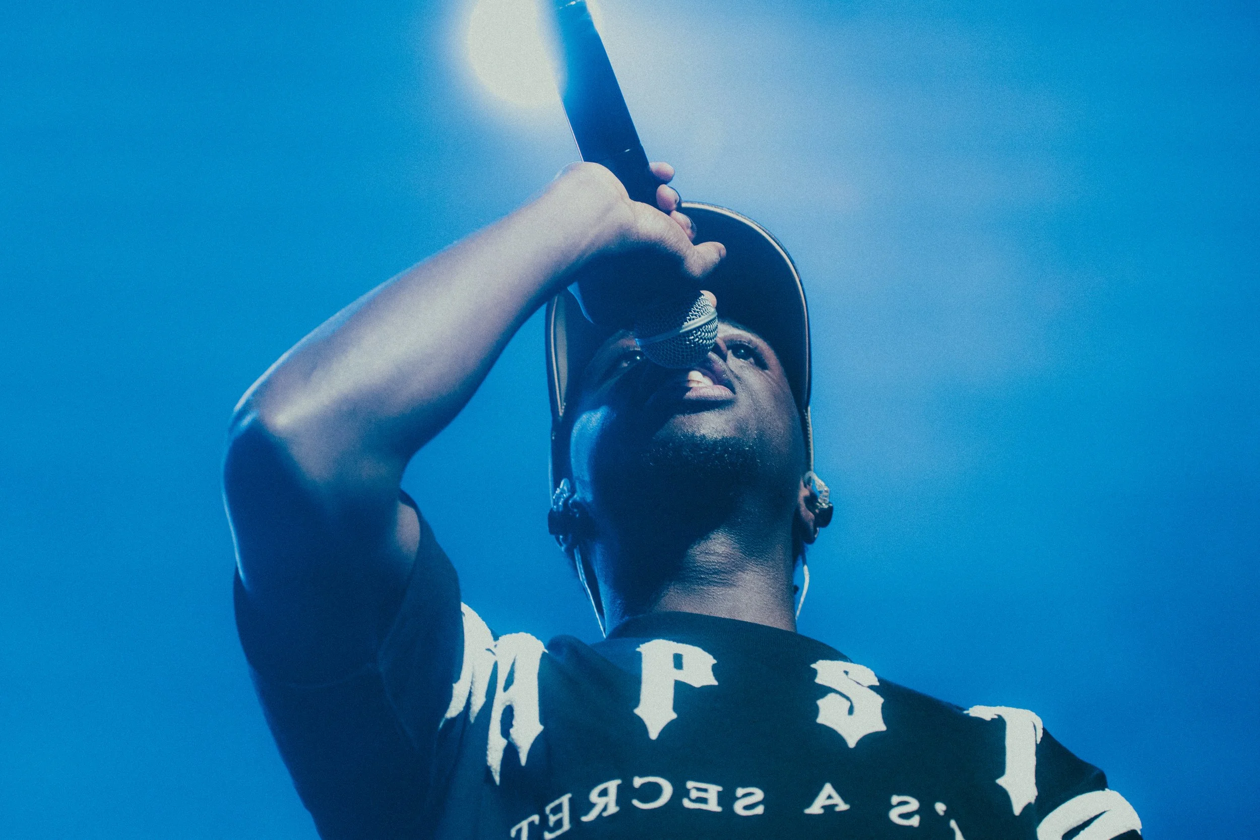 Un jeune homme portant un t-shirt noir avec des lettres blanches et une casquette, tenant un microphone et chantant ou parlant, avec un ciel bleu en arrière-plan.