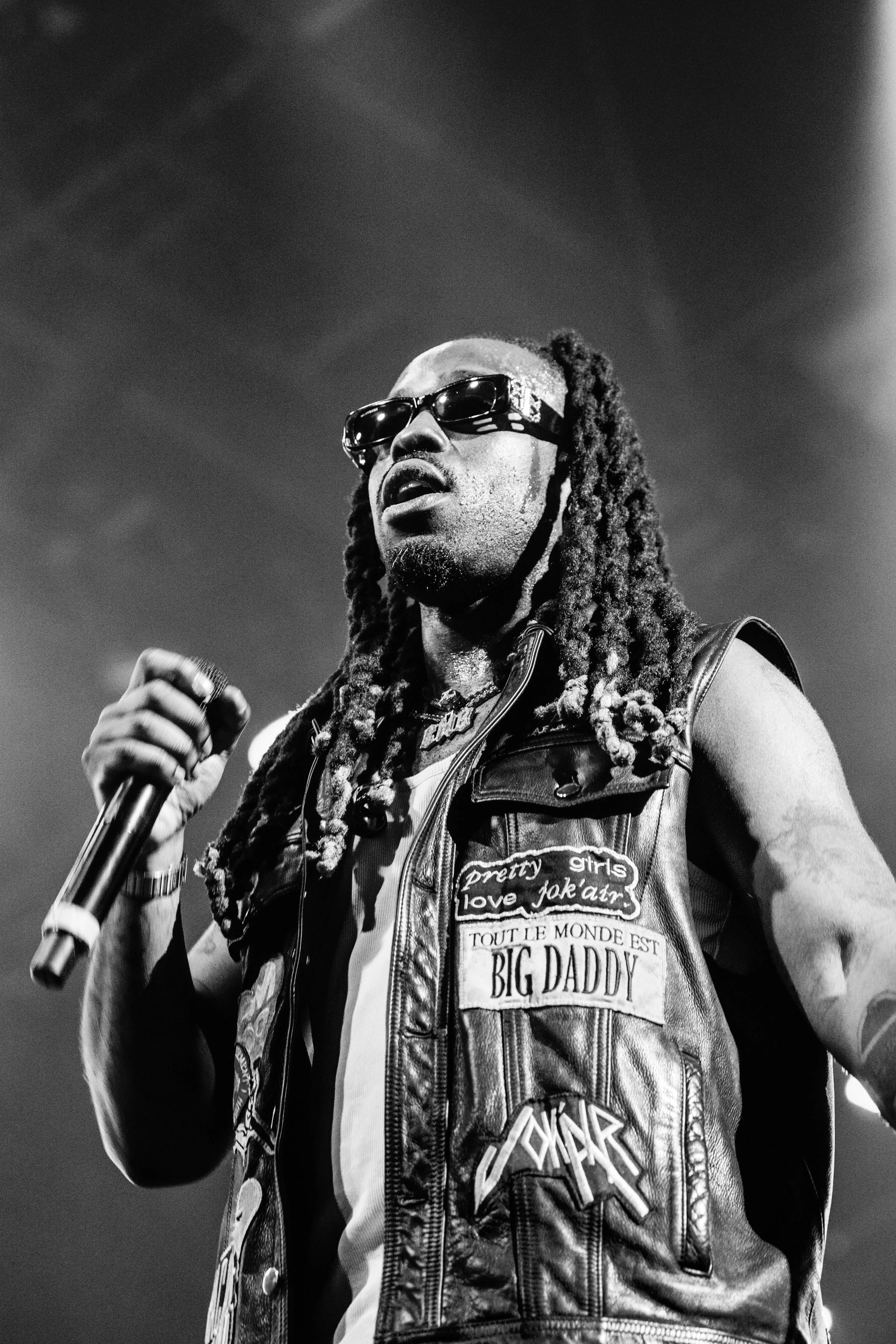 Un homme avec des dreadlocks, portant des lunettes de soleil et un gilet en cuir orné de plusieurs patchs, tient un micro lors d'une performance sur scène.