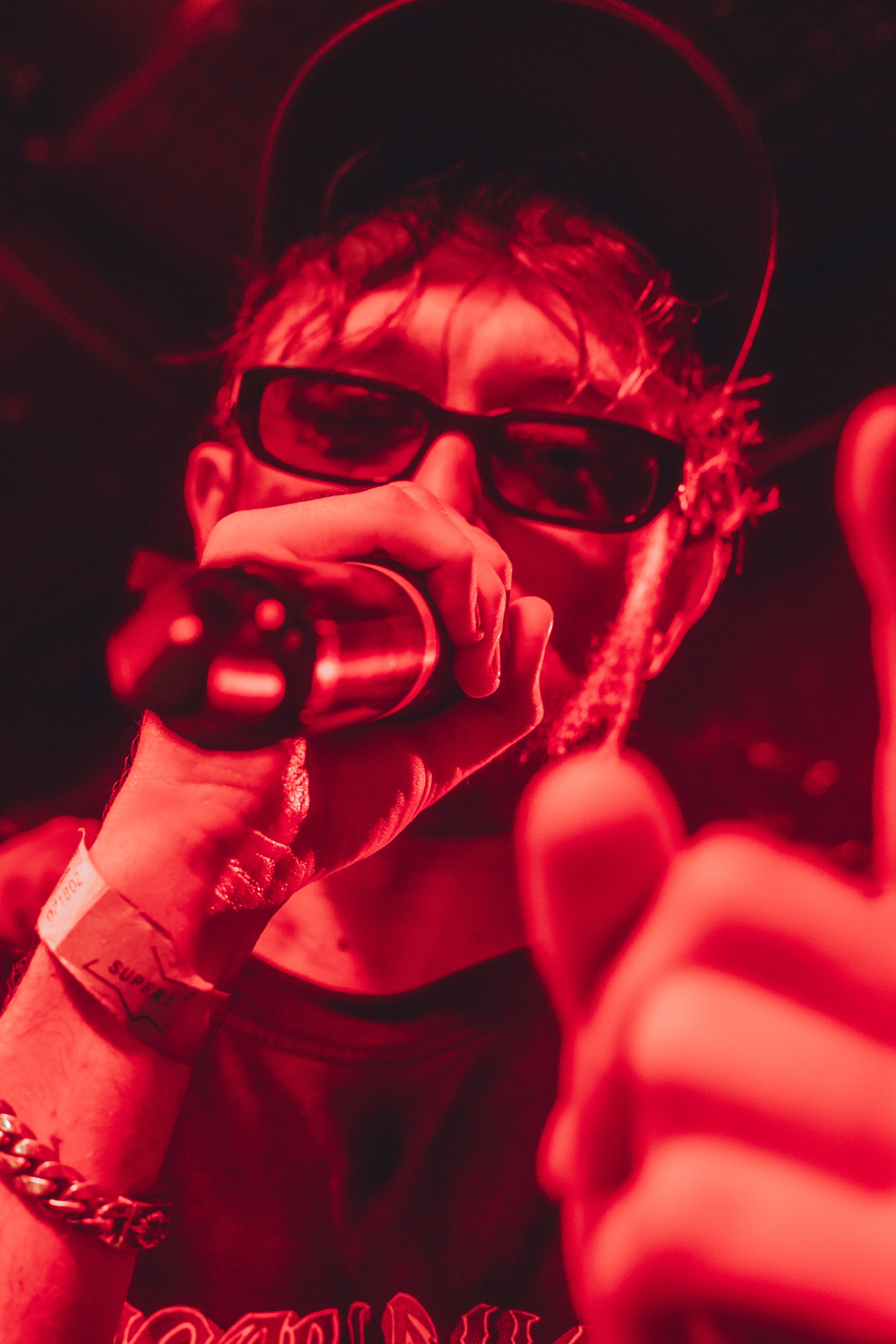 Une personne portant des lunettes de soleil noires et un casque, tenant un microphone, dans un environnement sombre avec un éclairage rouge intense.