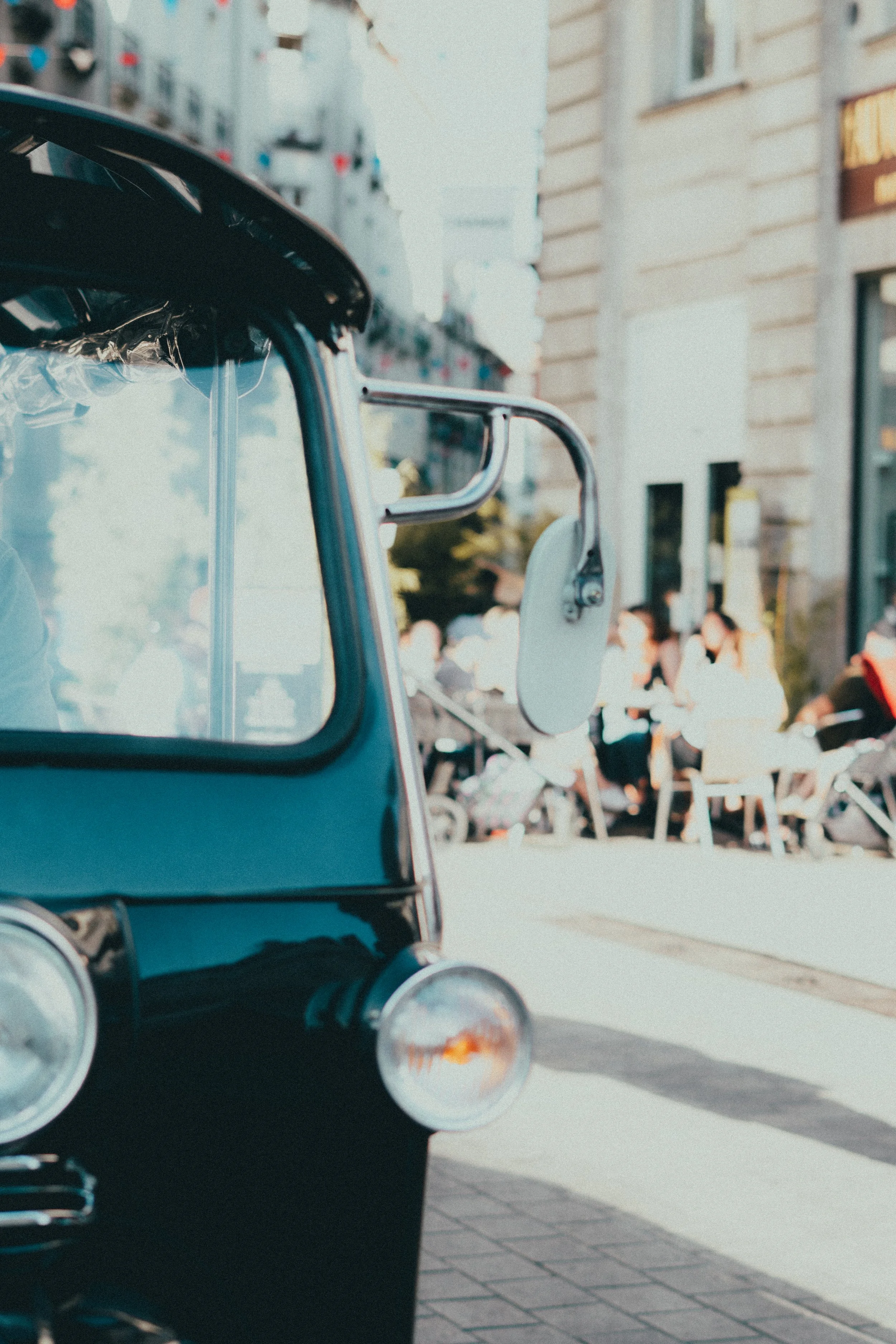 Une photo d'un véhicule à trois roues, probablement une rickshaw ou tuk-tuk, stationné sur un trottoir en ville, avec des personnes assises à une terrasse de café au fond.