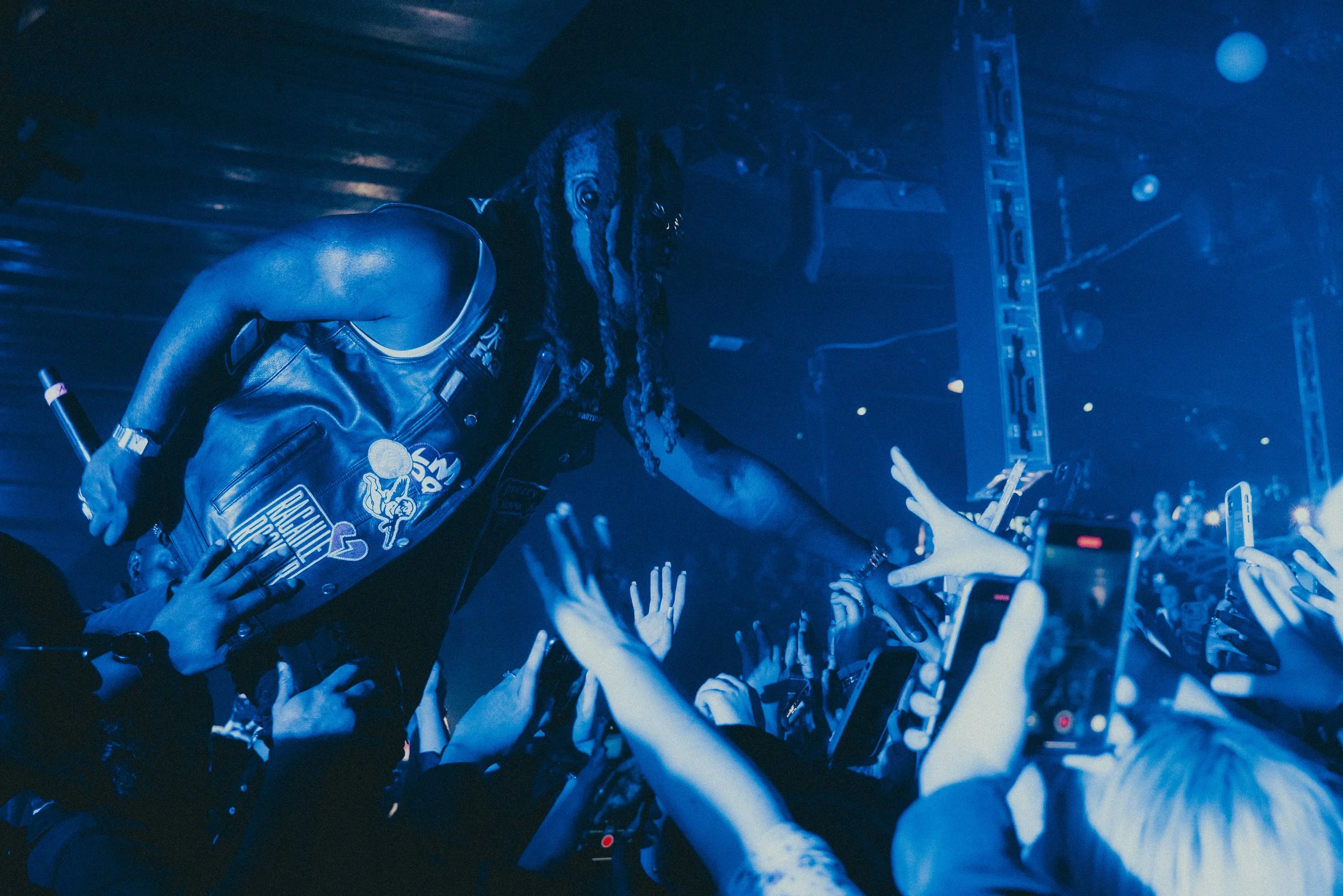 Un artiste avec des dreadlocks, portant un gilet noir orné de patches, tendant la main vers des fans lors d'un concert avec une ambiance sombre et éclairage bleu, nombreuses personnes en avant-garde avec des téléphones.