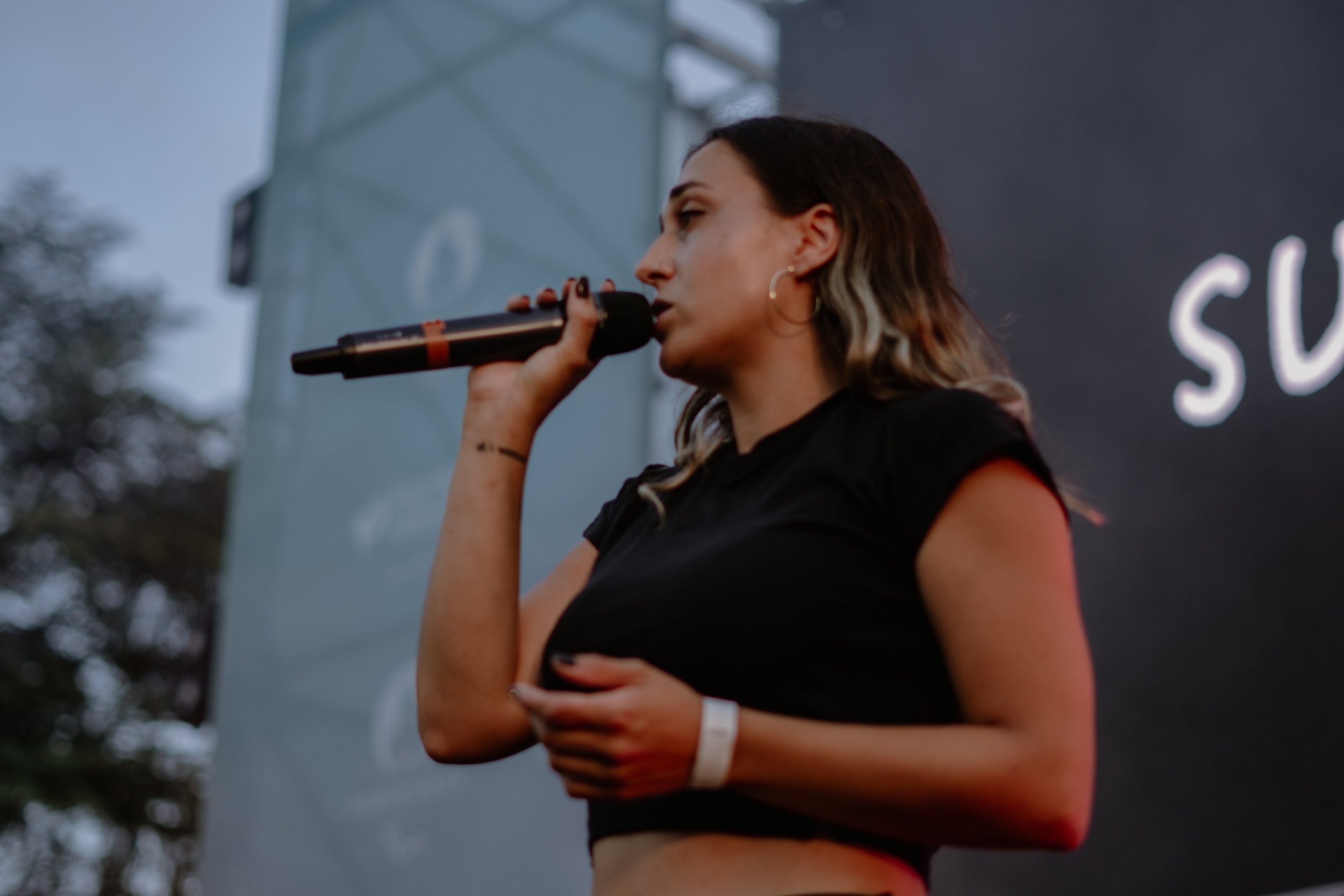 Une femme chante dans un microphone lors d'un événement en plein air, avec un fond sombre et des arbres en arrière-plan.