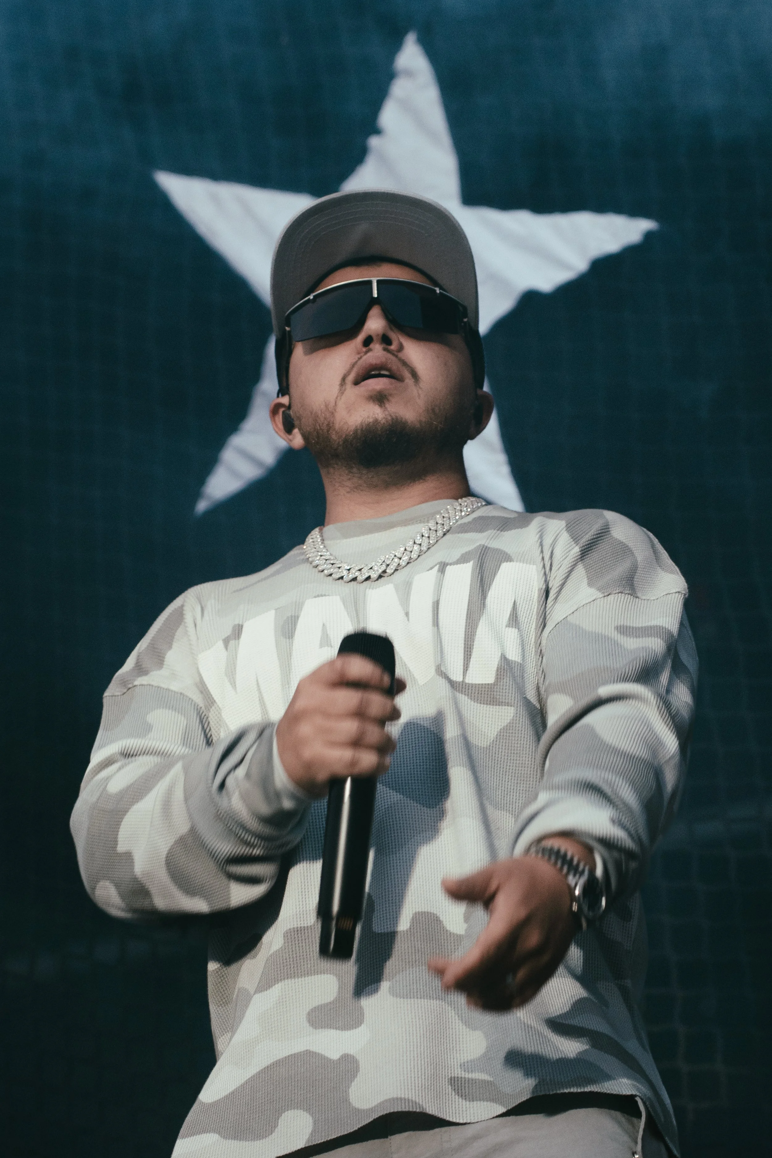 Un homme portant une casquette, des lunettes de soleil, un collier en chaîne, et un sweat à motif camouflage, tenant un microphone, avec un fond représentant une étoile blanche sur fond bleu.