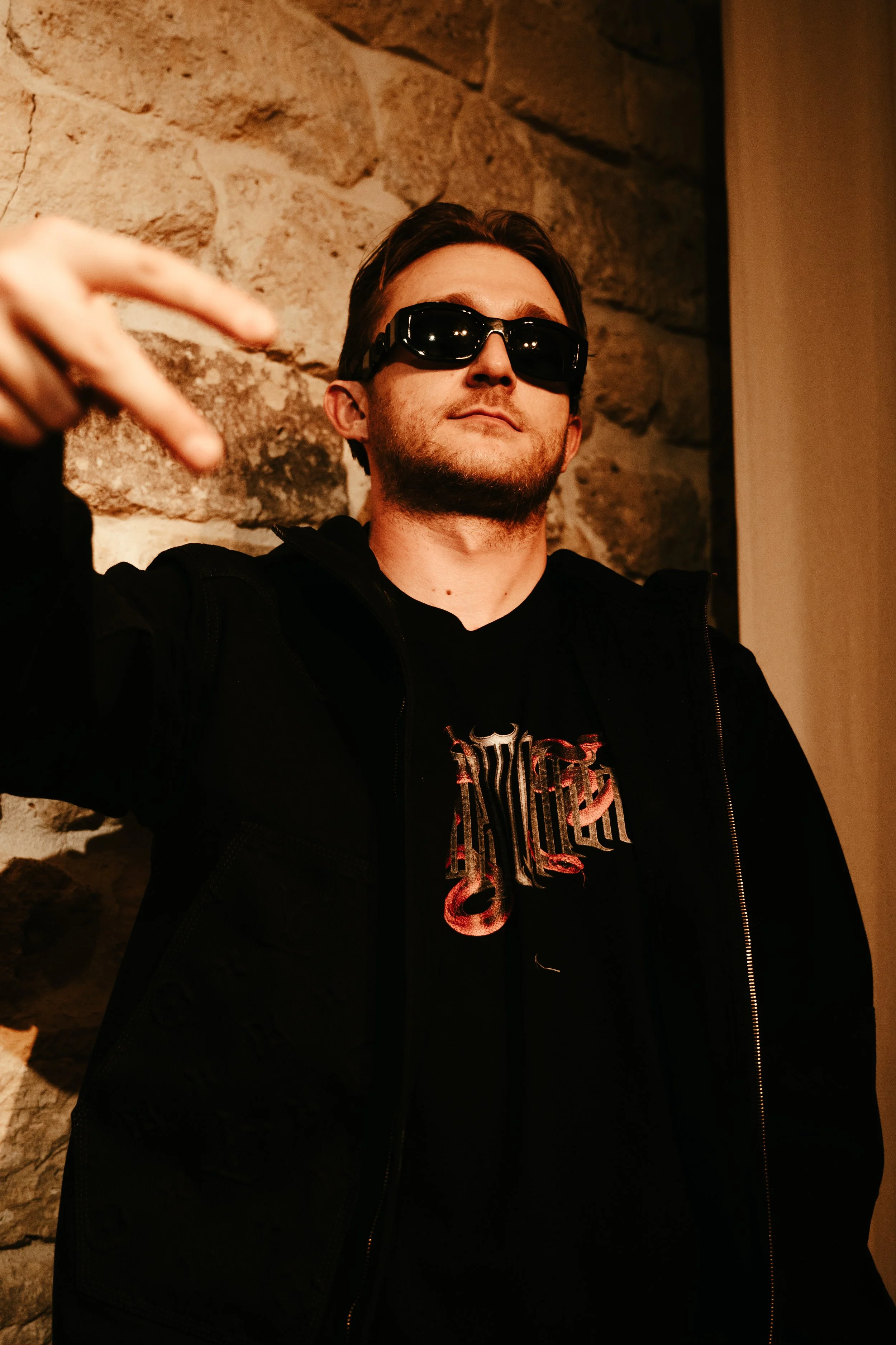 Jeune homme avec des lunettes de soleil noires, portant un hoodie noir, faisant un geste de paix avec la main devant un mur en pierre.