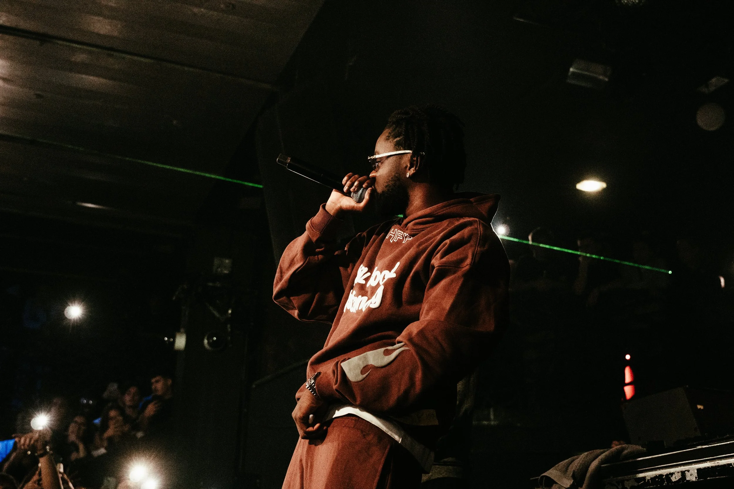 Un homme avec des dreads portant un sweat-brass marron et des lunettes, tenant un microphone, se produisant sur scène lors d'un concert, avec un public dans l'obscurité