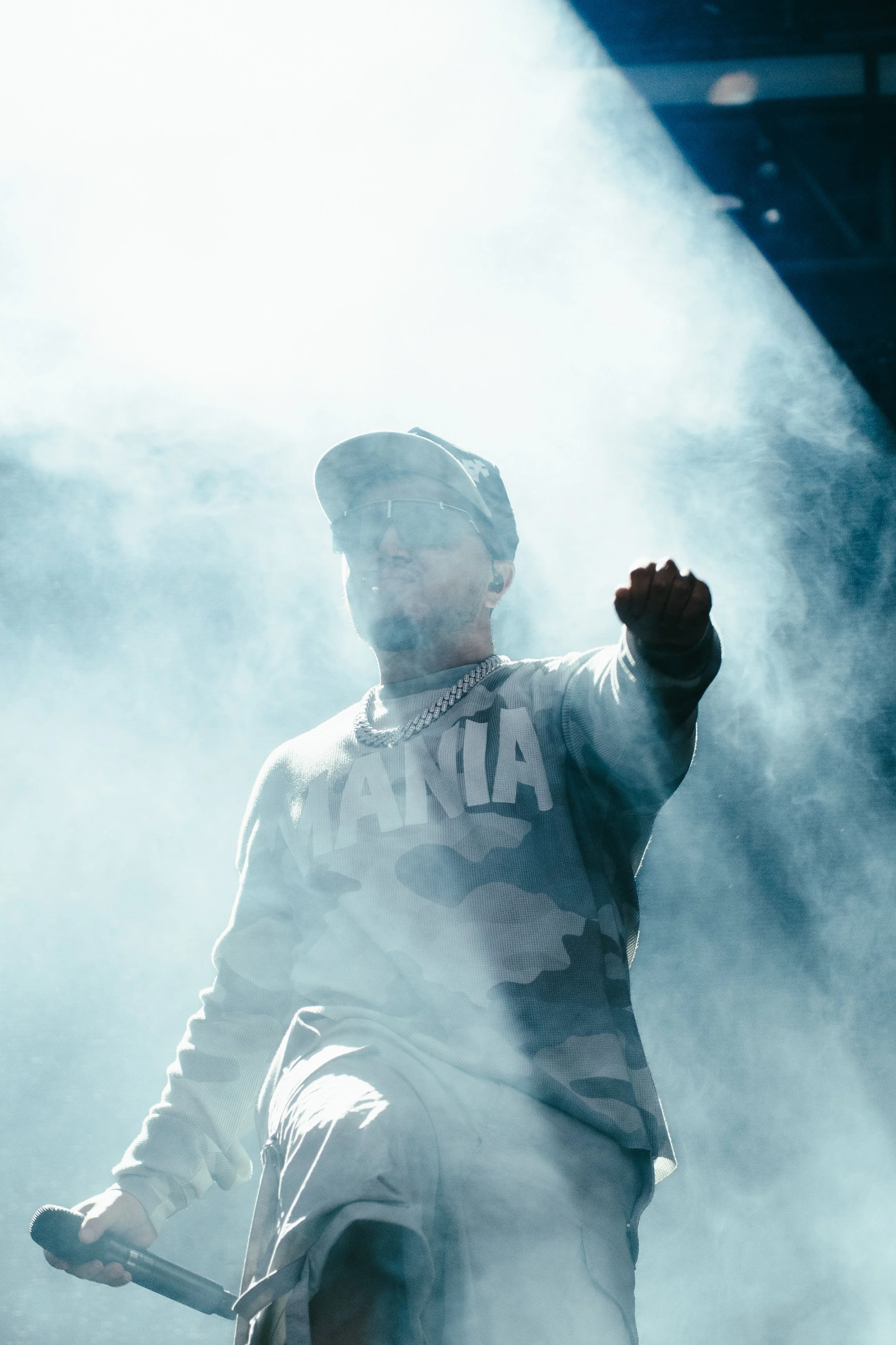 Un homme portant une casquette, des lunettes de soleil, un maillot avec le mot "MANIA" en lettre majuscule, une chaîne en or, tient un microphone dans la main et fait un geste de poing levé lors d'une performance sur scène avec un éclairage intense e