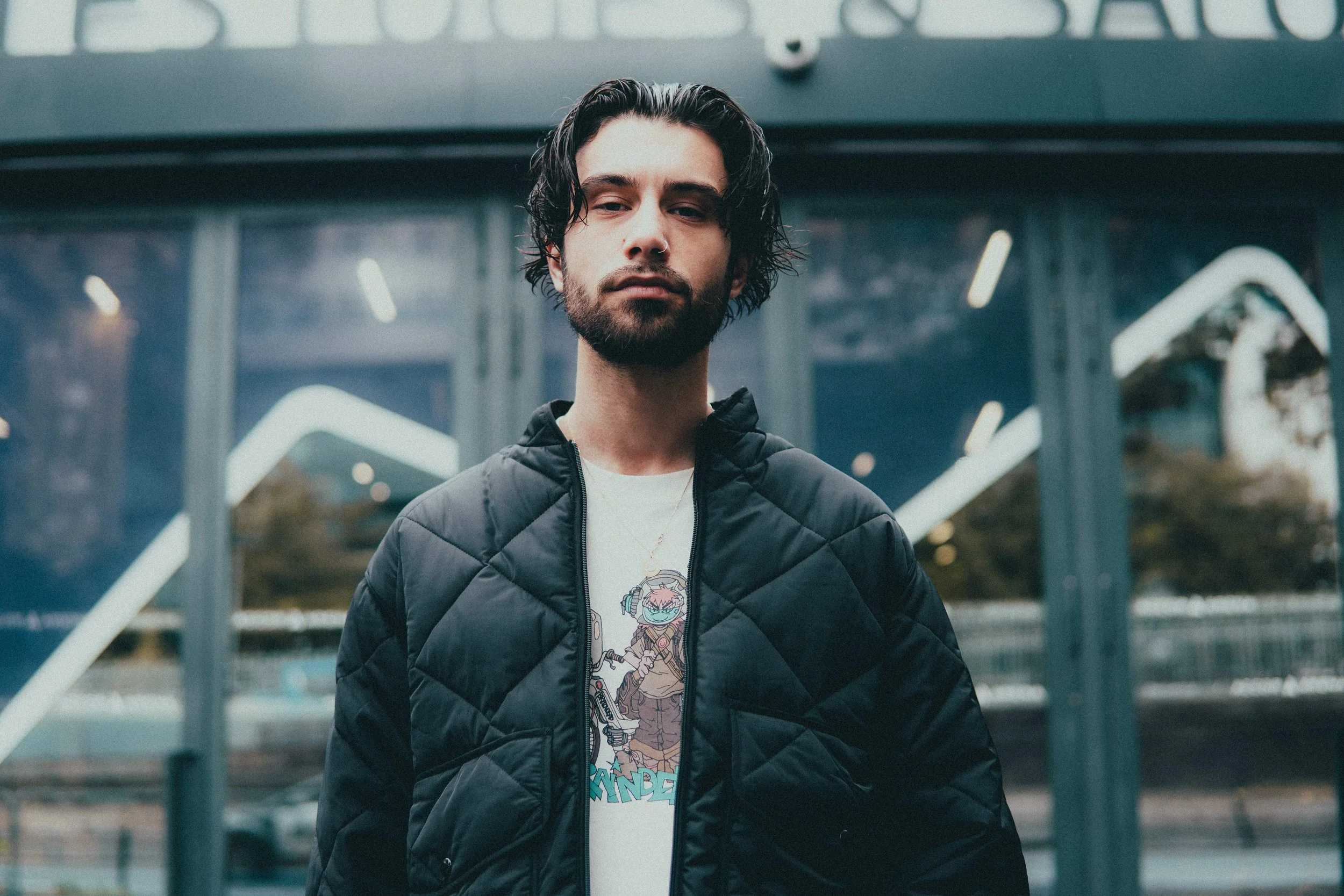 Un jeune homme avec des cheveux noirs bouclés, portant une veste noire matelassée et un t-shirt avec un dessin animé, se tient devant une structure de verre moderne en milieu urbain.