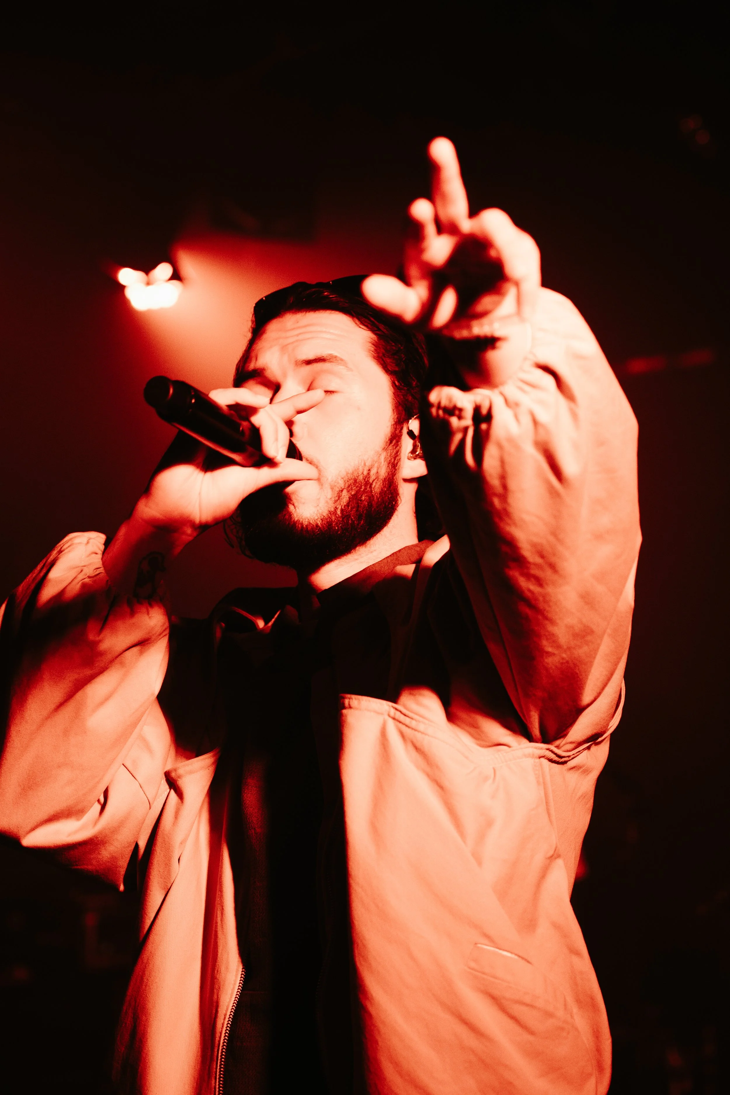 Un homme avec une barbe chante dans un micro, portant un blouson beige, sous un éclairage rouge intense.