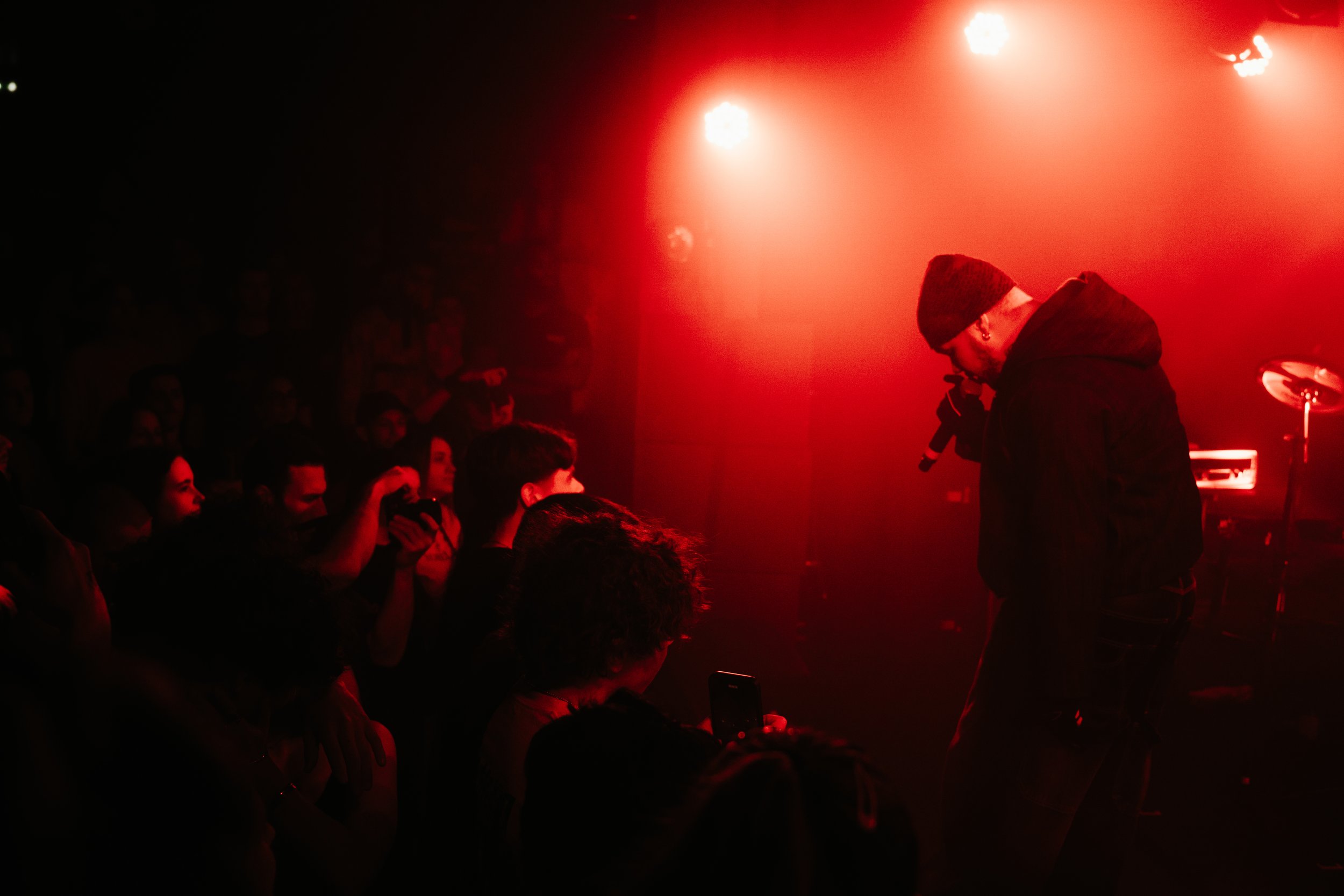 Un musicien ou chanteur avec un bonnet et une veste noire, tenant un microphone, sur scène dans une ambiance sombre et rouge, face à un public.