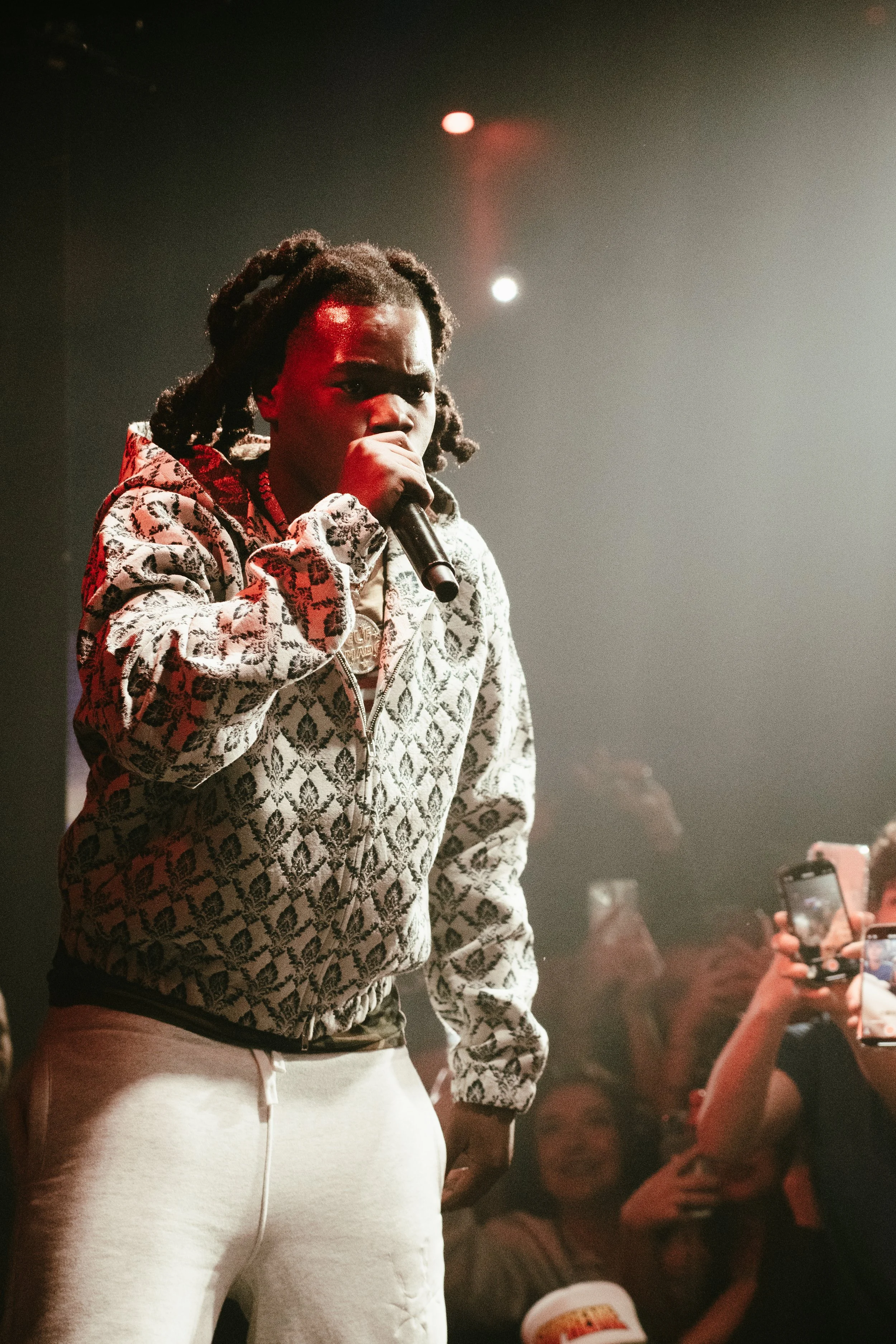 Jeune homme avec des dreadlocks tenant un microphone lors d'une performance, portant un sweat à motif, en arrière-plan un public enthousiaste.