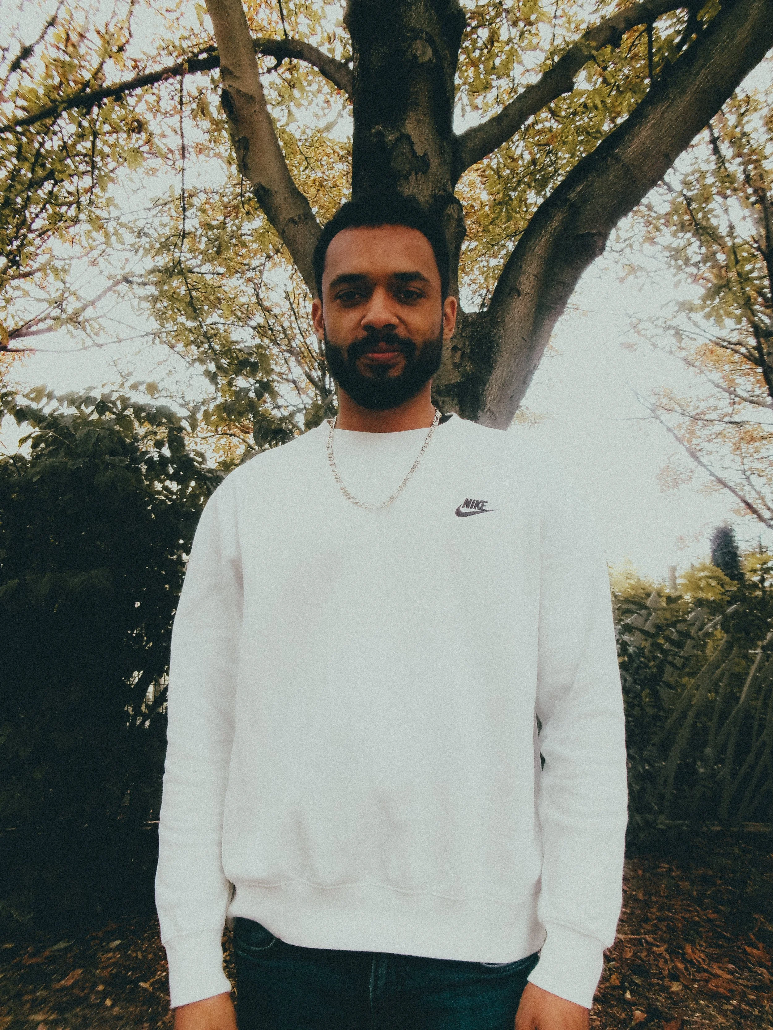 Un homme avec une barbe, portant un sweat blanc Nike, se tient devant un arbre avec des feuilles jaunes.