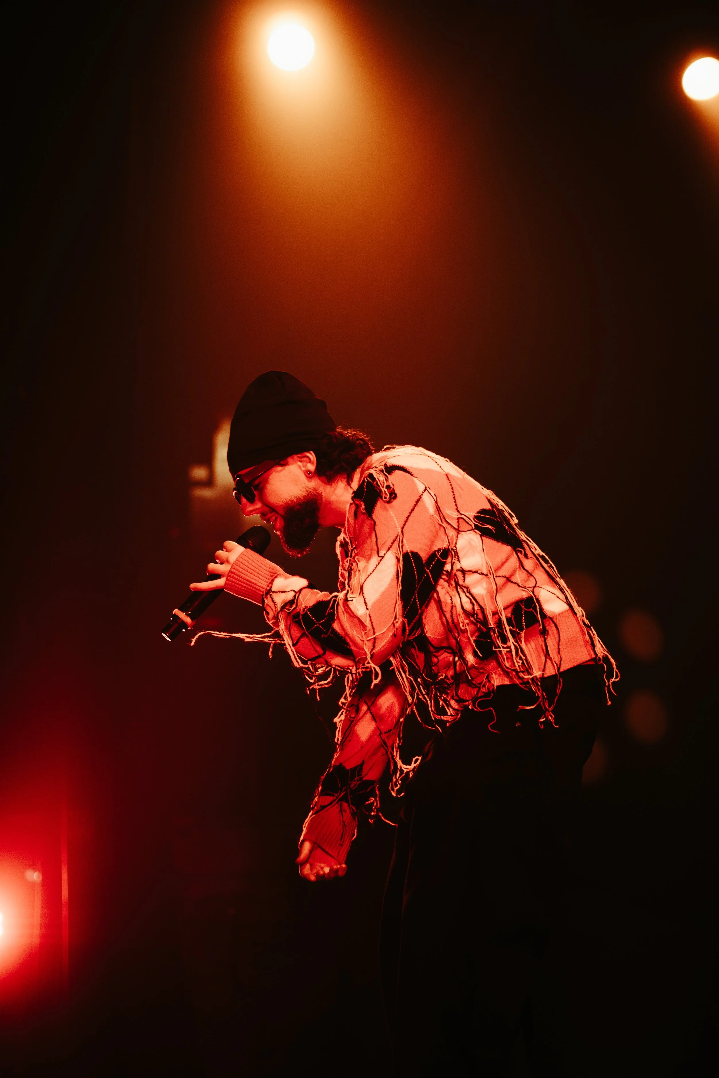 Un homme portant un bonnet noir, des lunettes de soleil, et un sweat déchiré, chante dans un microphone sur scène, sous un éclairage chaud et tamisé.