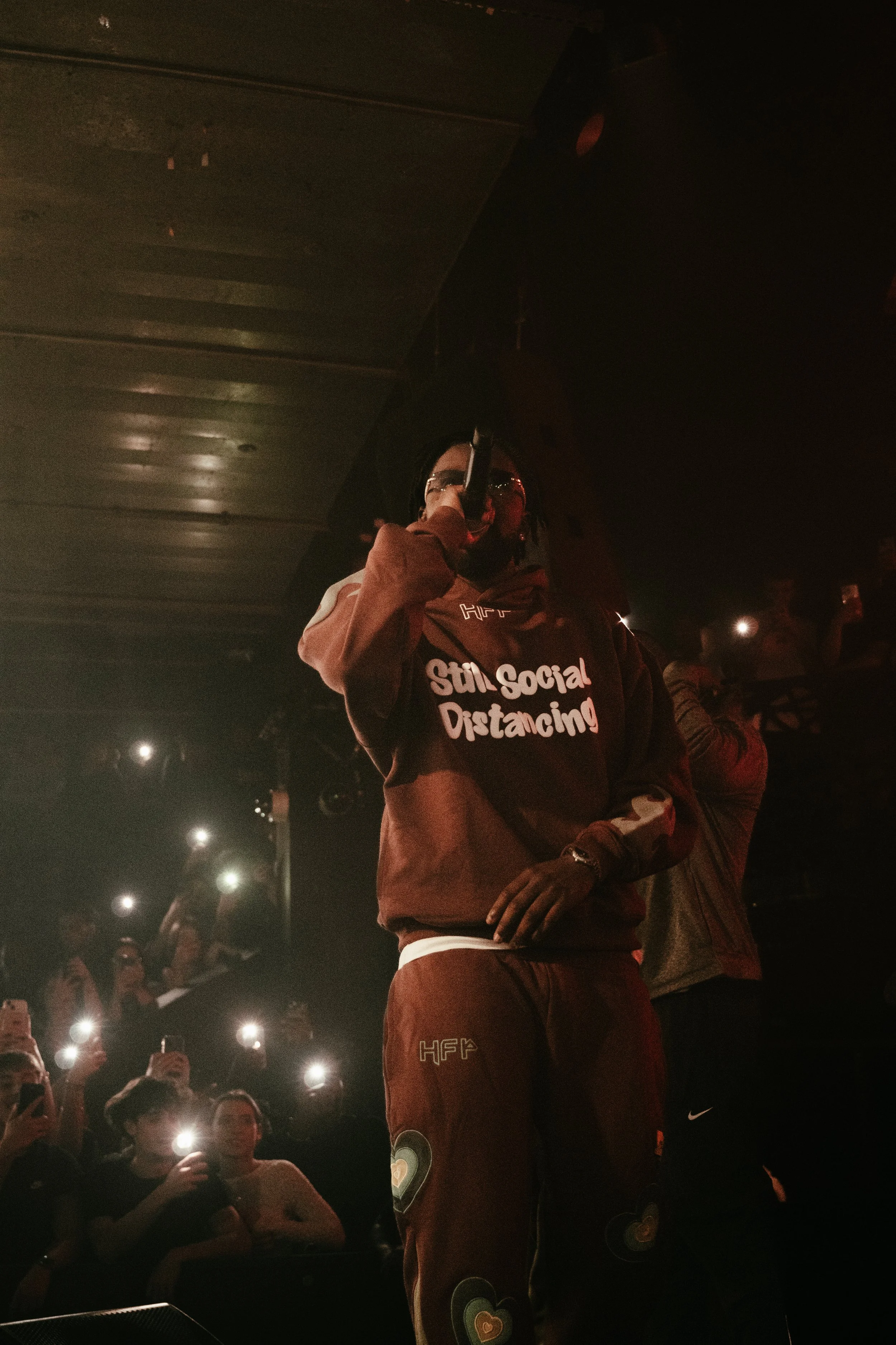 Un homme portant une veste avec le texte "Still Social Distancing" chante ou parle dans un micro sur scène, avec un public qui prend des photos avec leurs téléphones.