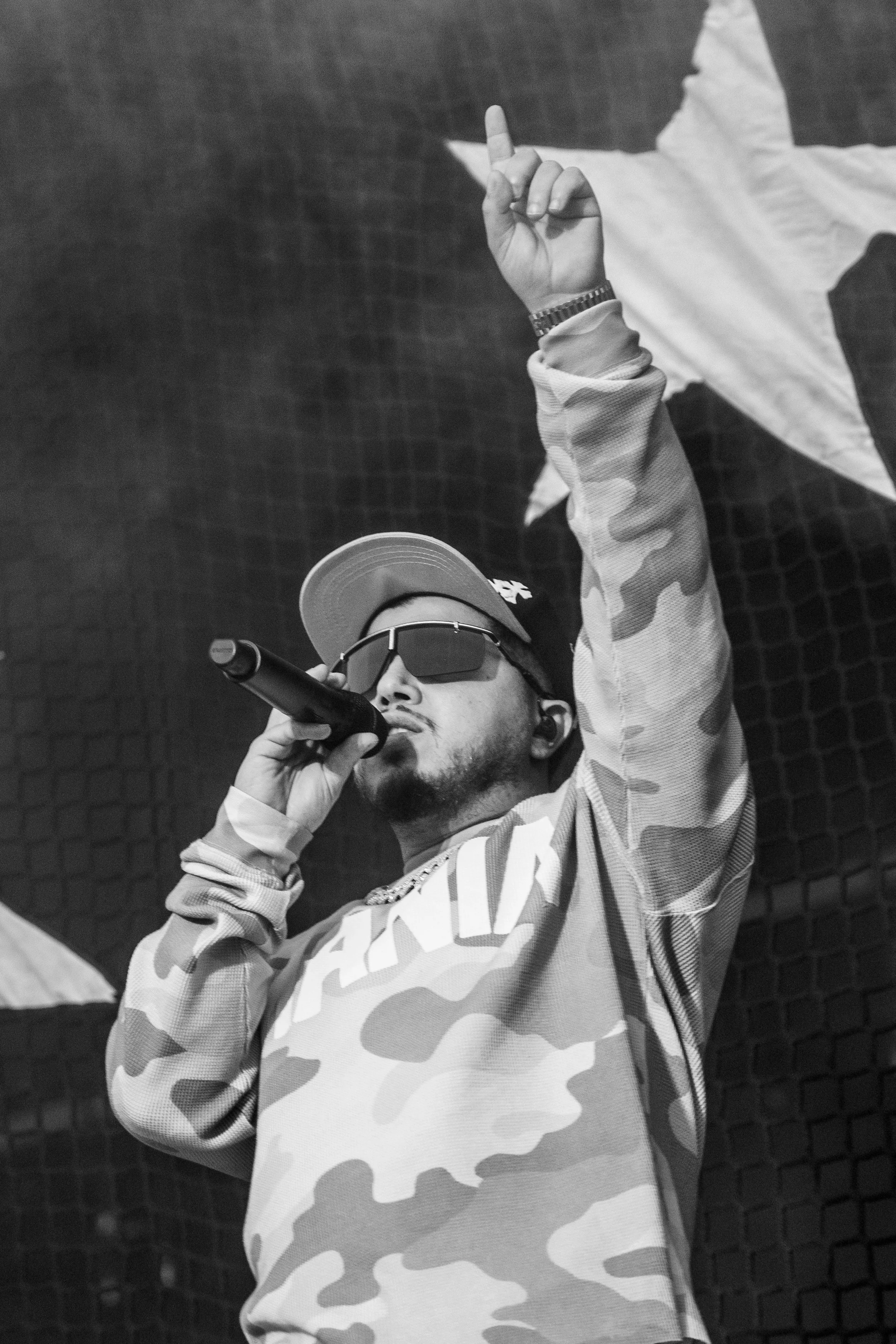 Un homme portant des lunettes de soleil, une casquette, un sweatshirt à motif camouflage, tenant un micro, et faisant un geste de la main avec le doigt levé.