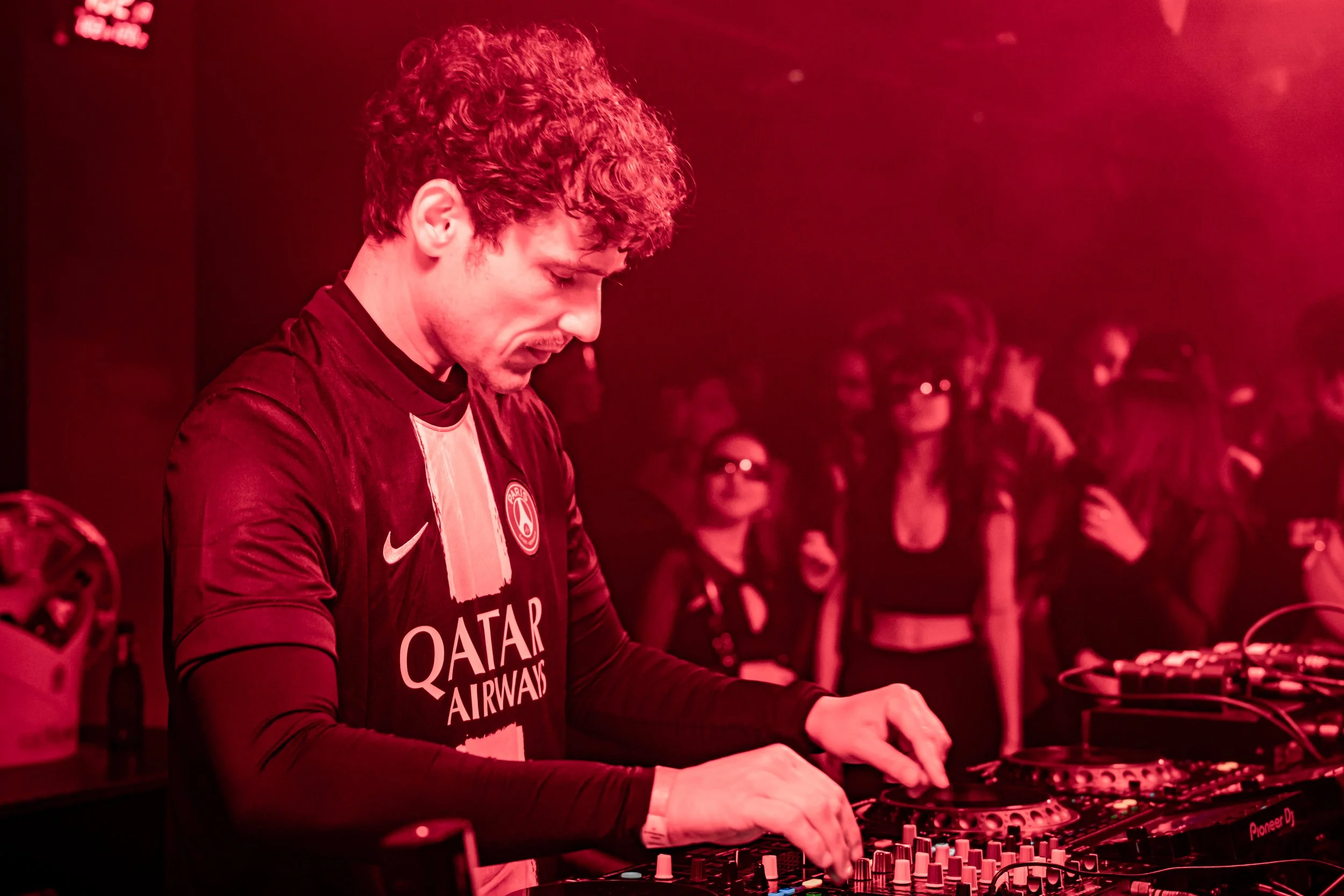Un DJ joue de la musique dans une boîte de nuit avec des personnes qui dansent en arrière-plan sous un éclairage rouge.