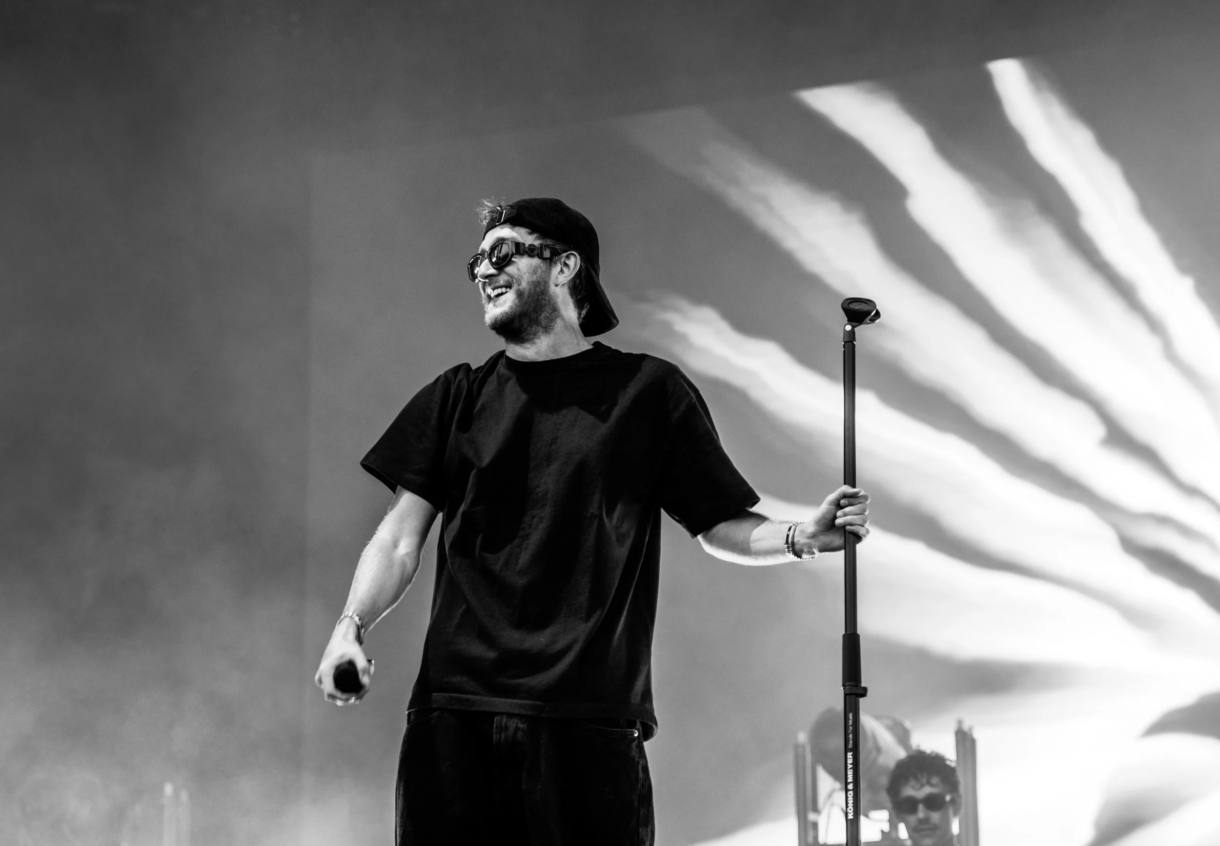 Un jeune homme souriant, portant des lunettes de soleil, un t-shirt noir, une casquette à l'envers, tenant un micro et un bâton de microphone, sur scène avec un fond de ciel et de nuages.
