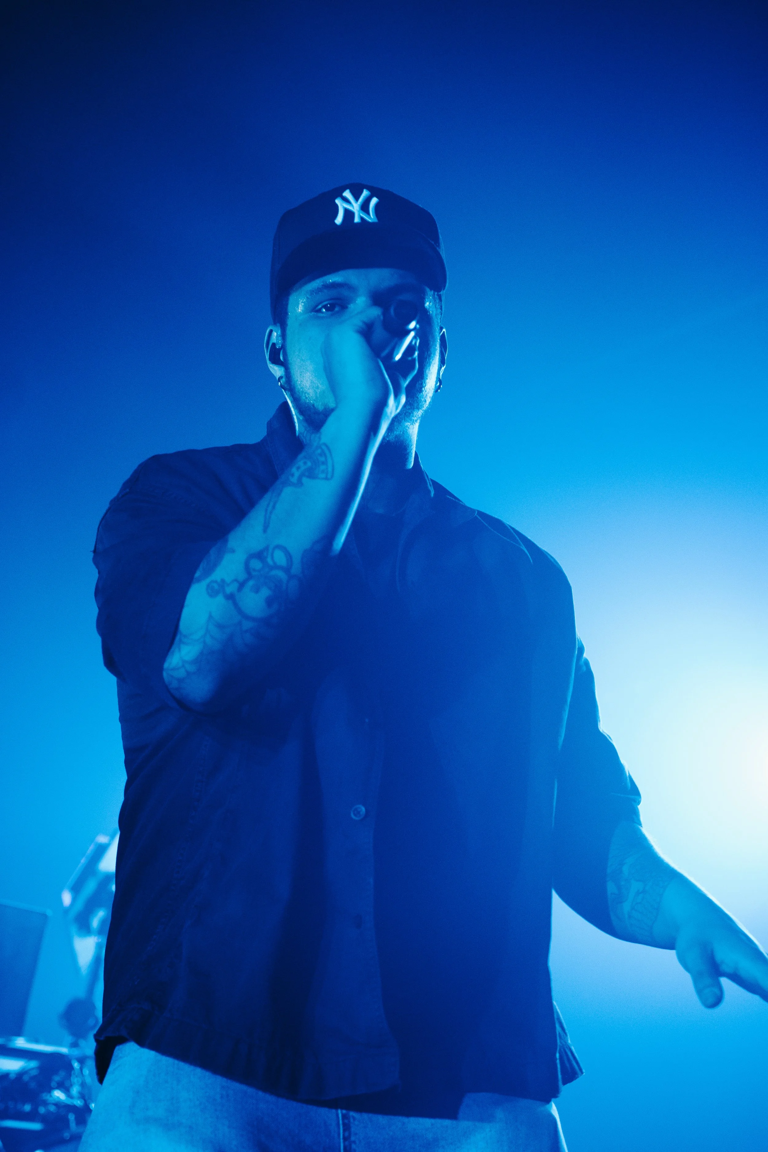 Un homme tenant un microphone en train de chanter sur scène, portant une casquette New York Yankees, avec un éclairage bleu et des tatouages visibles sur le bras.