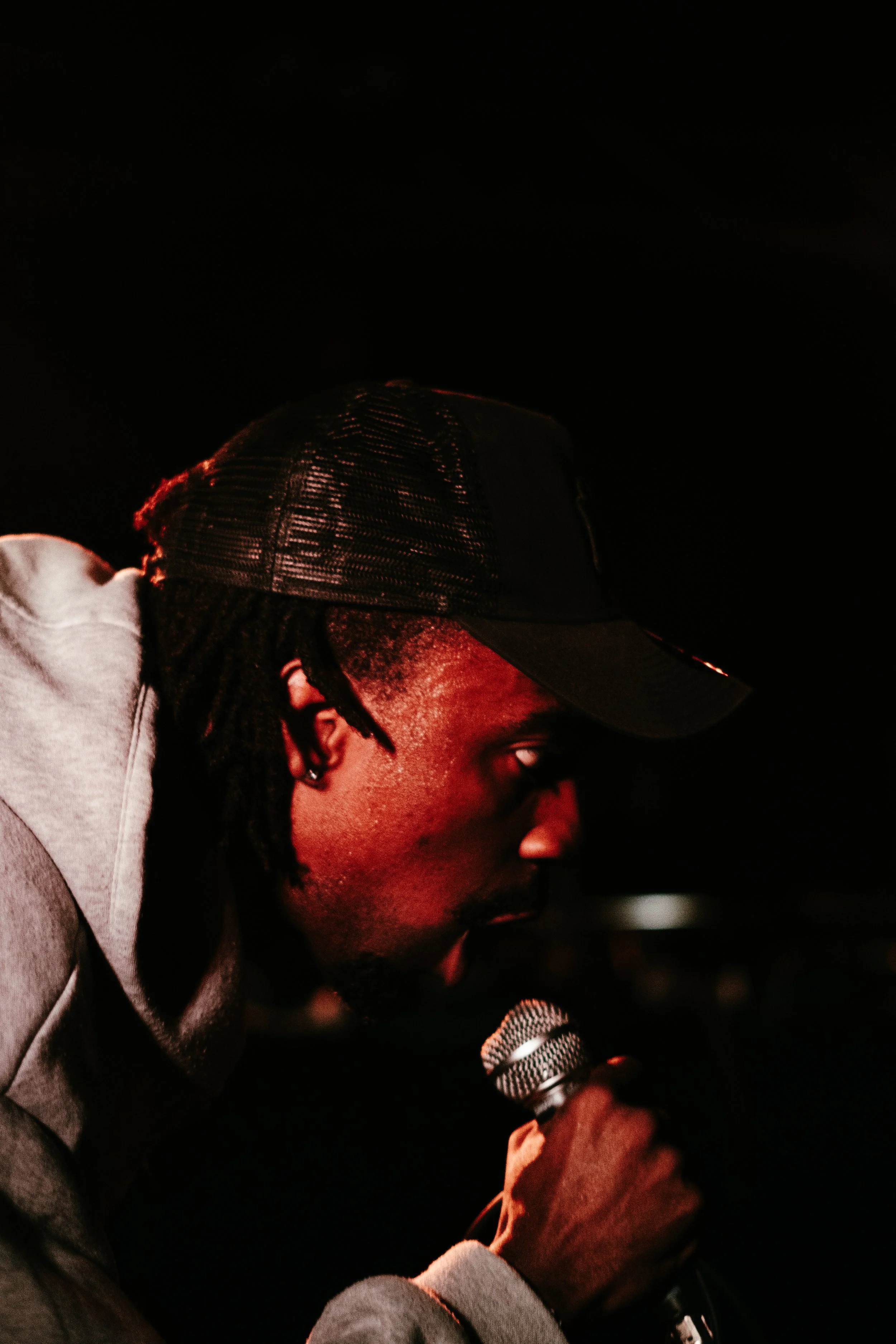 Jeune homme parlant dans un microphone, portant une casquette noire et un sweat gris, dans un cadre sombre