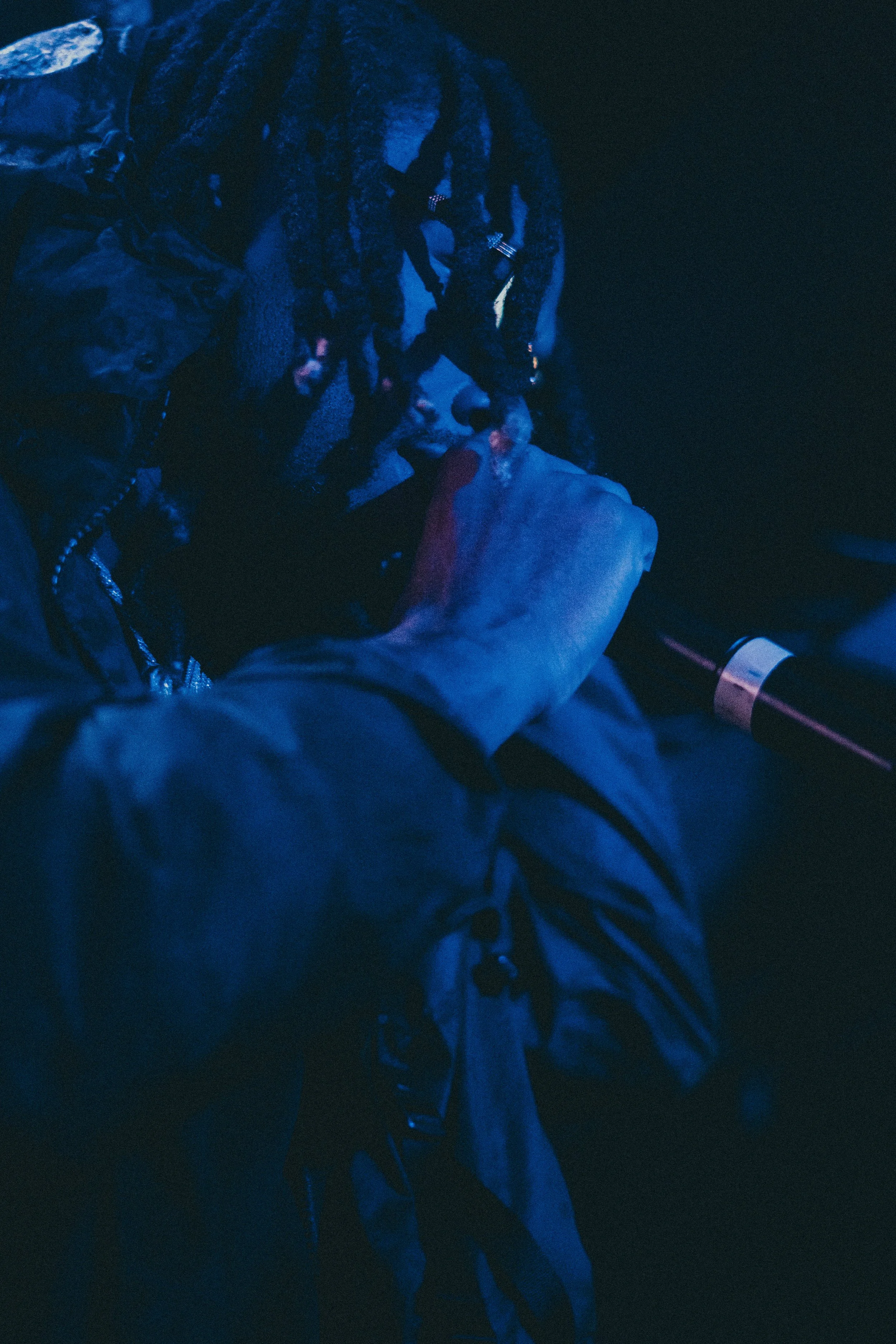 Un homme les yeux fermés, en tenue sombre, souffle dans une longue pipe ou tube, dans un environnement sombre avec un éclairage bleu.