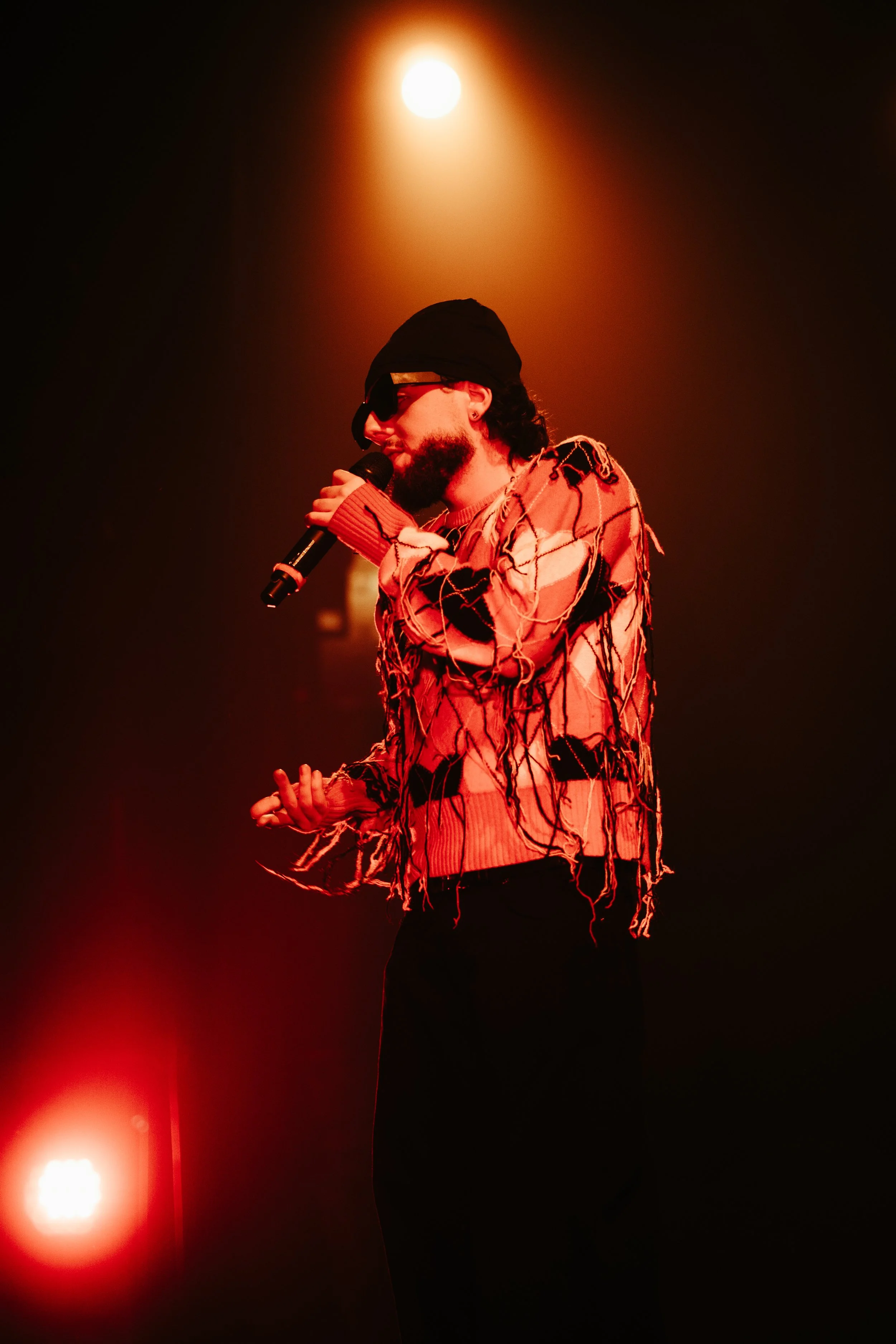 Un homme portant un bonnet noir, des lunettes de soleil et un sweat-shirt déchiré et tissé, chantant dans un microphone sur scène avec un éclairage chaud en arrière-plan.