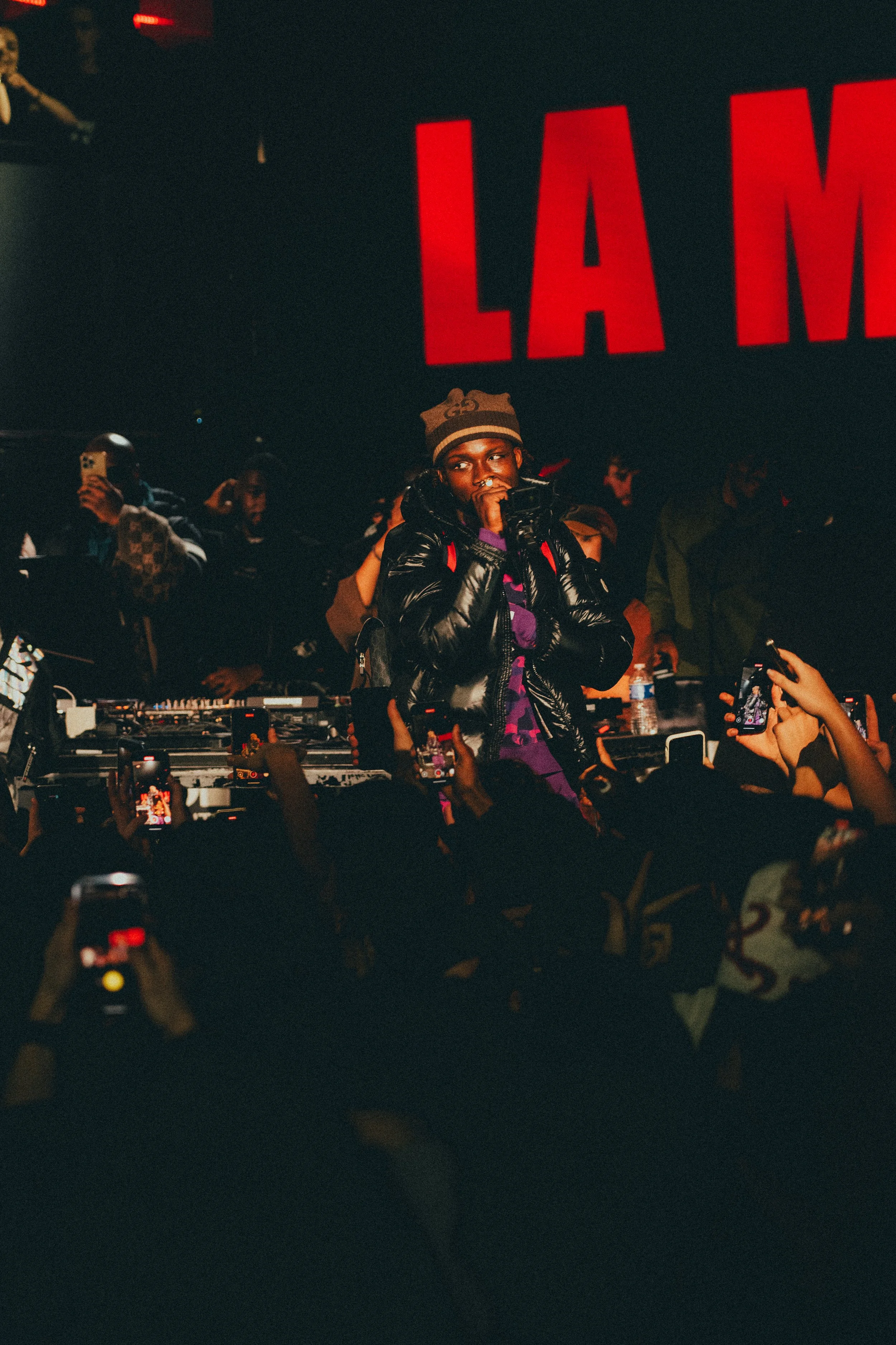 Un artiste ou DJ en train de performer sur scène lors d'un concert ou d'un événement musical, entouré d'un public pris de photos ou vidéos avec leur téléphone. Le fond comporte un grand écran avec des lettres rouges."