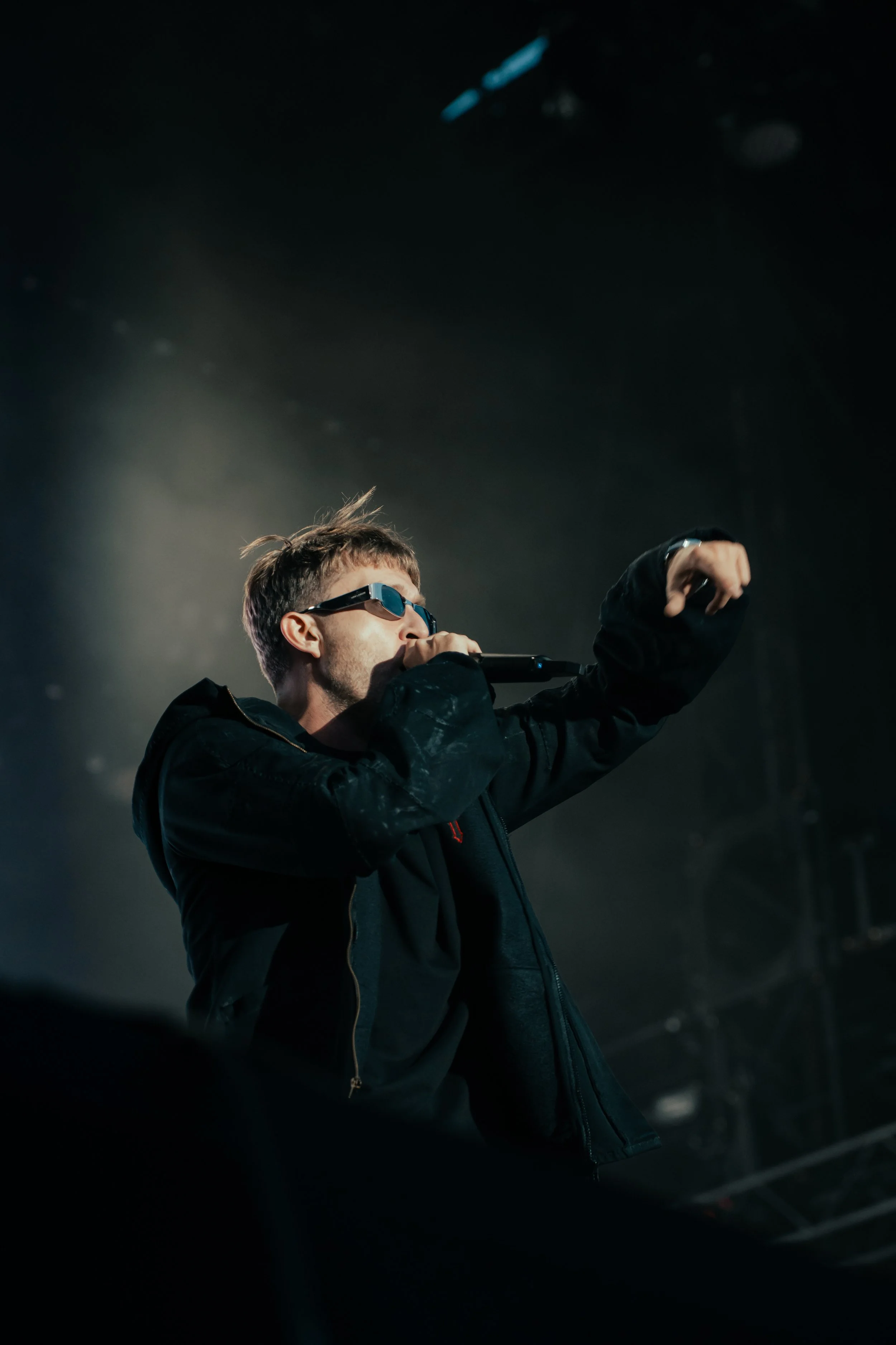 Un homme portant des lunettes de soleil noires chante dans un micro, habillé en noir, sur scène, sous des lumières tamisées.