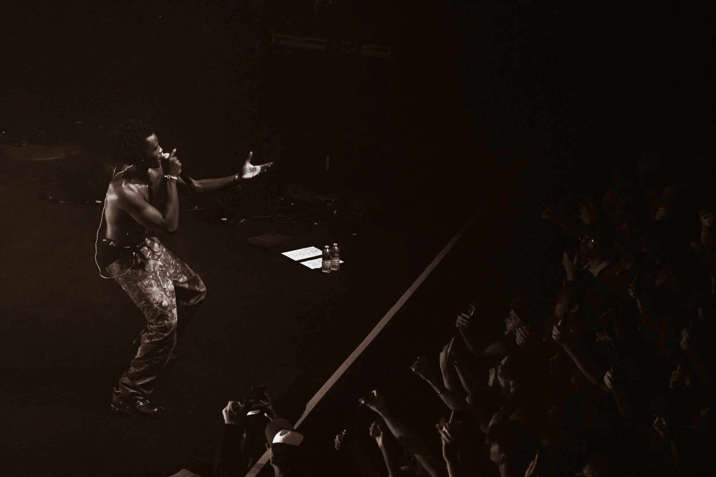 Un homme nu en train de parler ou chanter à une foule dans une salle de concert sombre, avec des bouteilles d'eau et des papiers sur le sol près de lui.