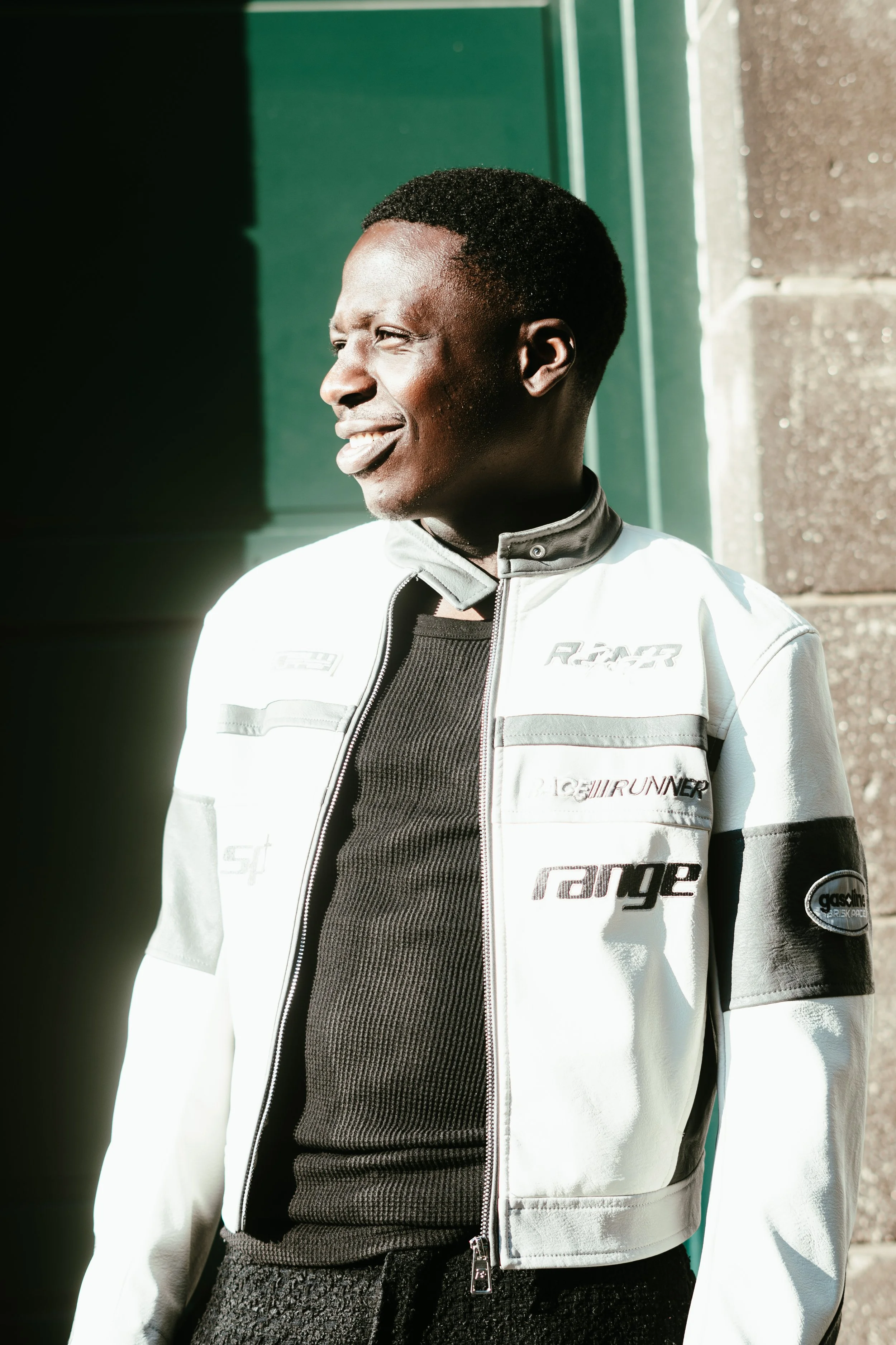 Jeune homme souriant portant une veste de motard blanche avec des logos, debout devant un bâtiment, dans la lumière du soleil.