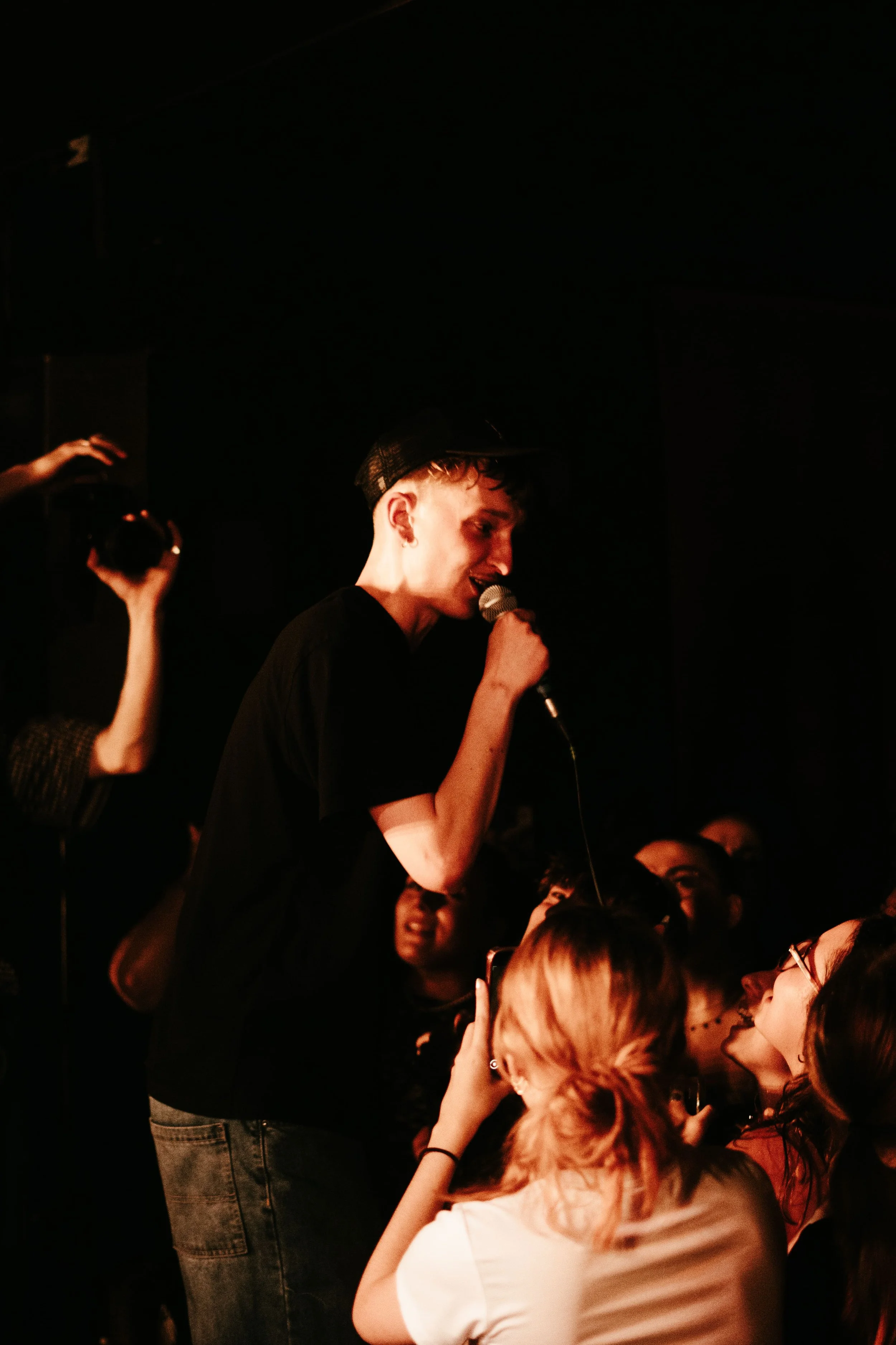 Un jeune homme chante avec un micro devant un groupe d'admirateurs lors d'une performance dans une salle sombre.