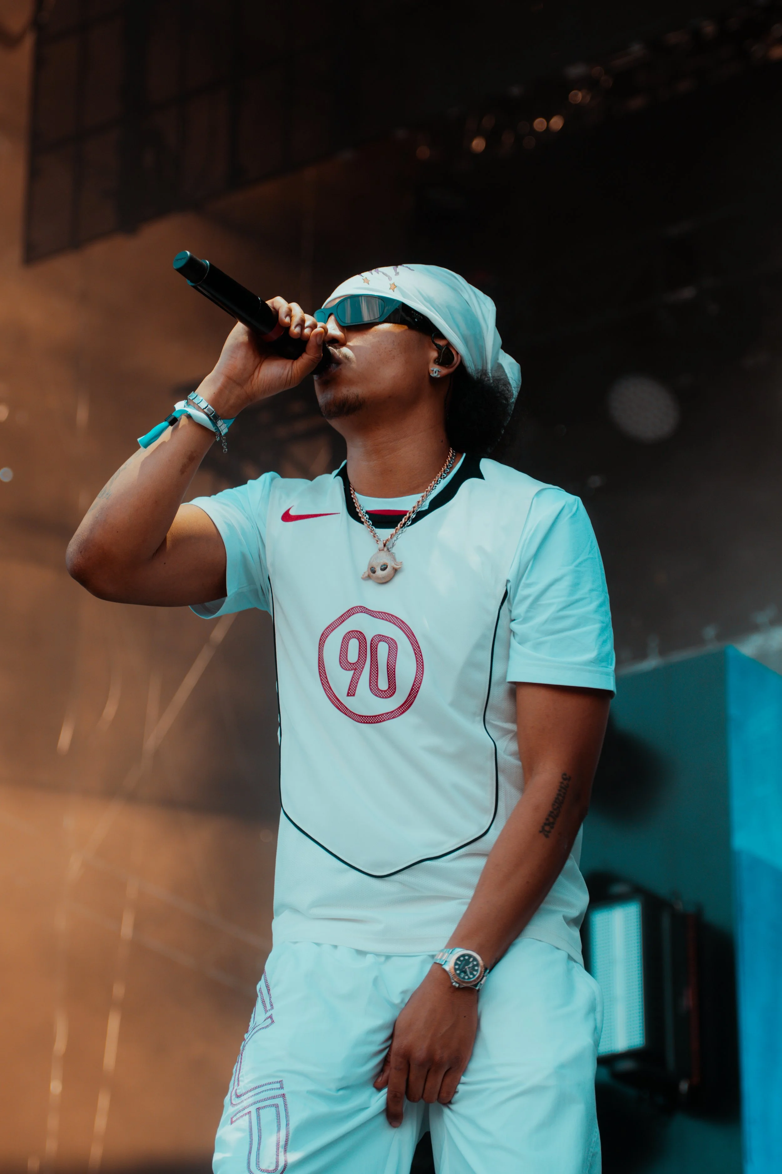 Une personne en tenue blanche, portant des lunettes de soleil et un bonnet, chante dans un microphone sur scène.